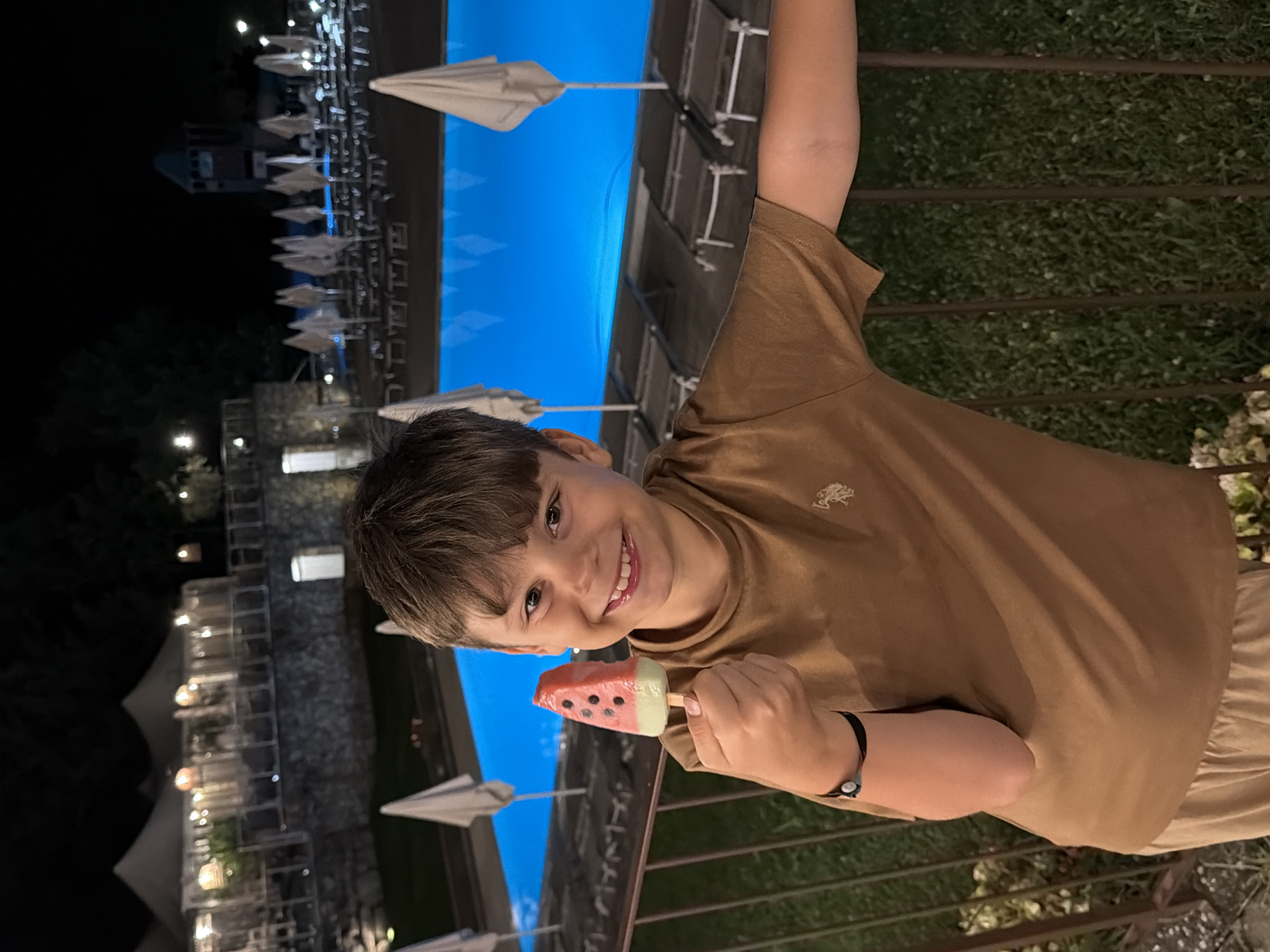 A young boy smiles while holding a watermelon-shaped popsicle in front of a brightly lit swimming pool at night. The boy is wearing a brown t-shirt and light-colored pants. In the background, there are lounge chairs, umbrellas, and buildings with lights. The setting appears to be a resort or hotel in Cavriglia, Italy.