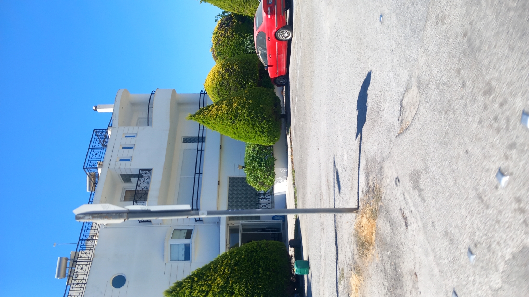 In the city of Lambi, Greece, an exterior scene is captured on a clear, sunny day with a bright blue sky. A white, multi-story building with balconies and solar water heaters on the rooftop dominates the background. In front of the building, a red sedan is parked. Several meticulously manicured evergreen trees and bushes are planted in front of the building. The foreground consists of a paved area, likely a street or parking lot, with some scattered debris and reflective markers on the surface. A traffic signpost is partially visible in the middle ground. A green plastic container is situated on the ground near the left side of the frame.