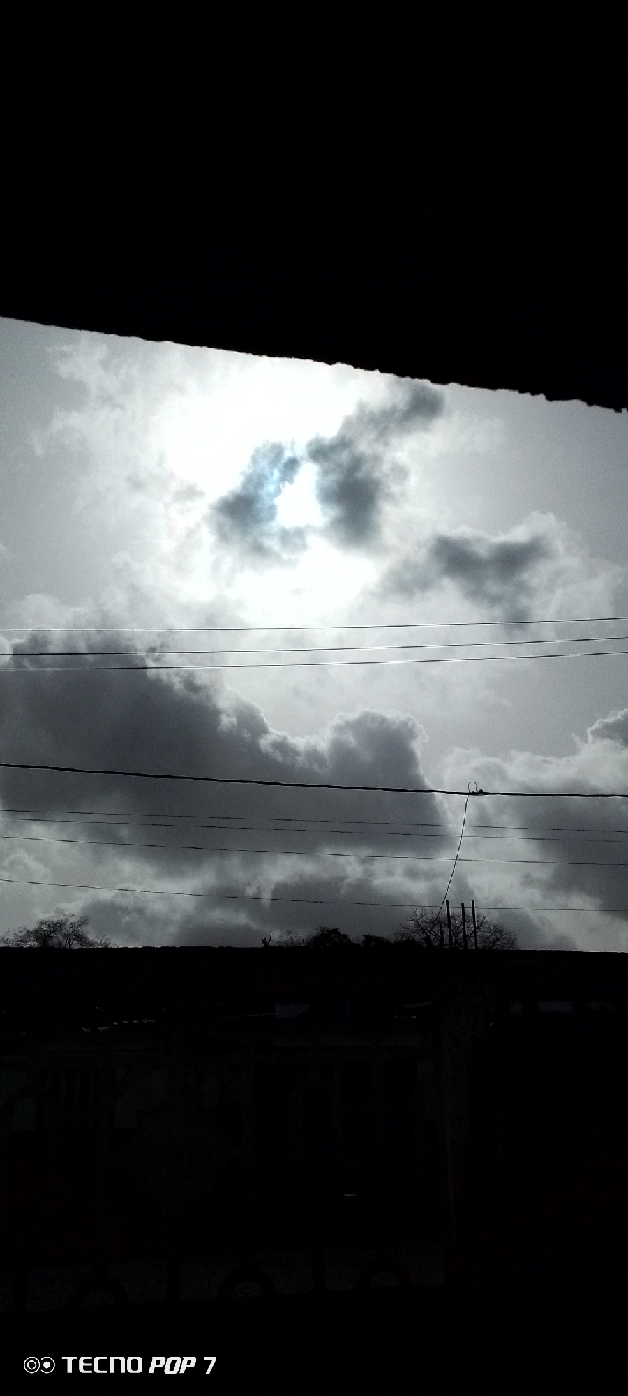 The image shows a view of a cloudy sky in Altagracia, Venezuela, framed by a dark structure, possibly a roof or building overhang. The sky is filled with grey and white clouds, with a brighter area indicating the sun. Several horizontal power lines stretch across the scene. Silhouetted trees and buildings are visible in the middle ground. A smartphone's camera watermark appears at the bottom.