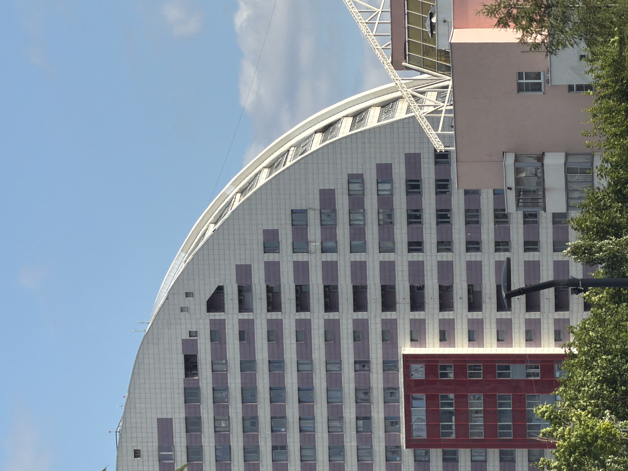 This media file depicts a modern architectural design featuring a curved building with a light-colored facade punctuated by rows of purple rectangles. The building has numerous windows, suggesting it is an office building or residential complex. The facade appears to be made of tiled panels, with a metallic sheen on some sections. A portion of a pink building with a different architectural style is visible to the right, also featuring windows. A red rectangular section is attached to the lower part of the curved building. Lush green trees partially obscure the view of the buildings, indicating an outdoor setting. A dark pole with a curved metal attachment extends into the frame from the right, and a white structural element, possibly part of a crane or scaffolding, is visible in the upper right quadrant. The sky is clear and blue, suggesting daytime with fair weather. The image is oriented vertically. There is no visible text in the media file.