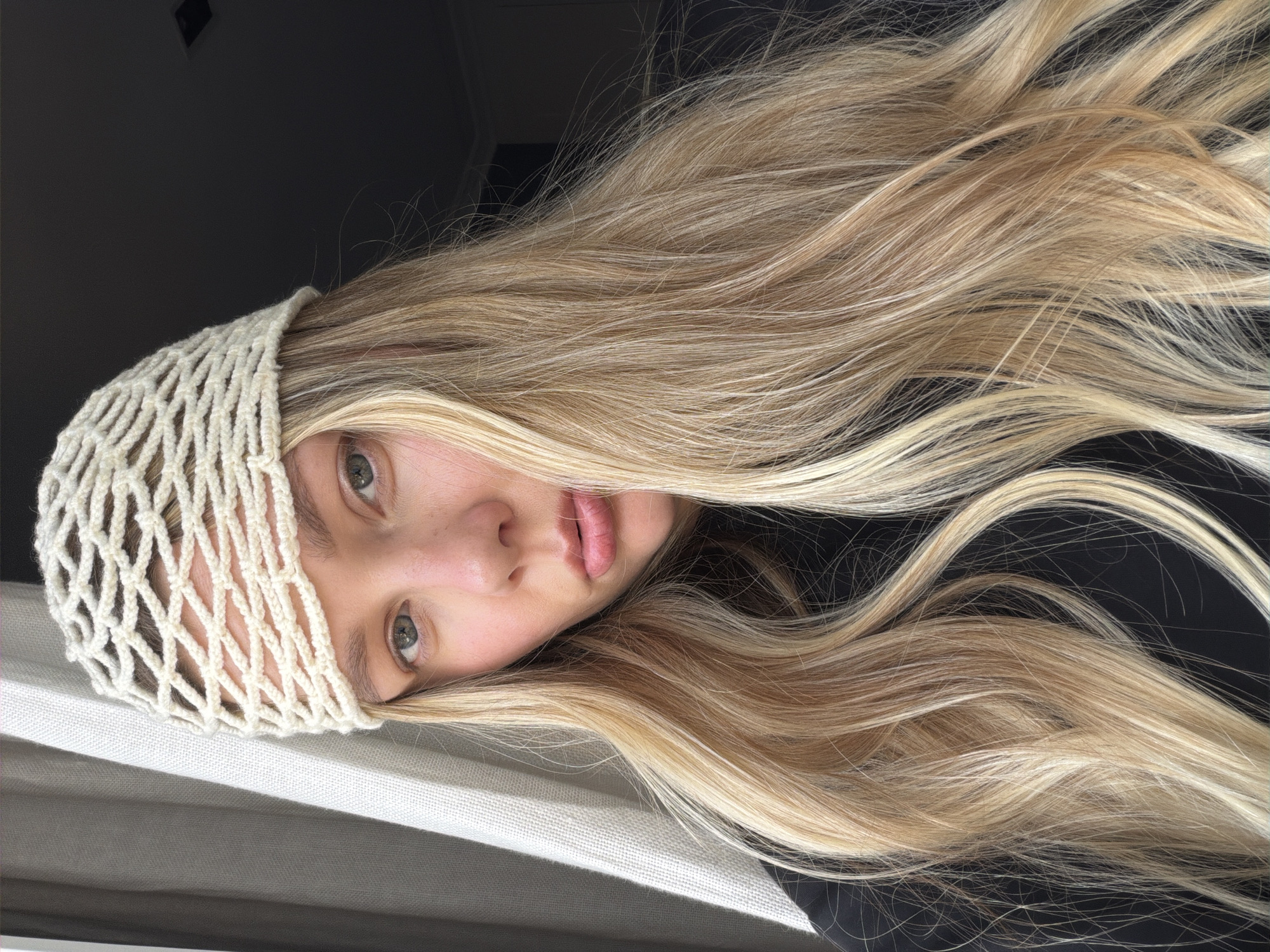 A close-up, rotated image shows a young person with long, blonde hair wearing a cream-colored knitted net-like beanie. The subject's face is partially visible, with one eye and lips in focus. The background is dark, with some lighter fabric or surface in the lower left quadrant, showing horizontal lines. The lighting suggests an indoor setting, possibly near a window, with shadows casting across the subject and the background. The subject's expression is neutral. There is no visible text. The image's orientation is sideways, with the subject's head tilted to the right.
