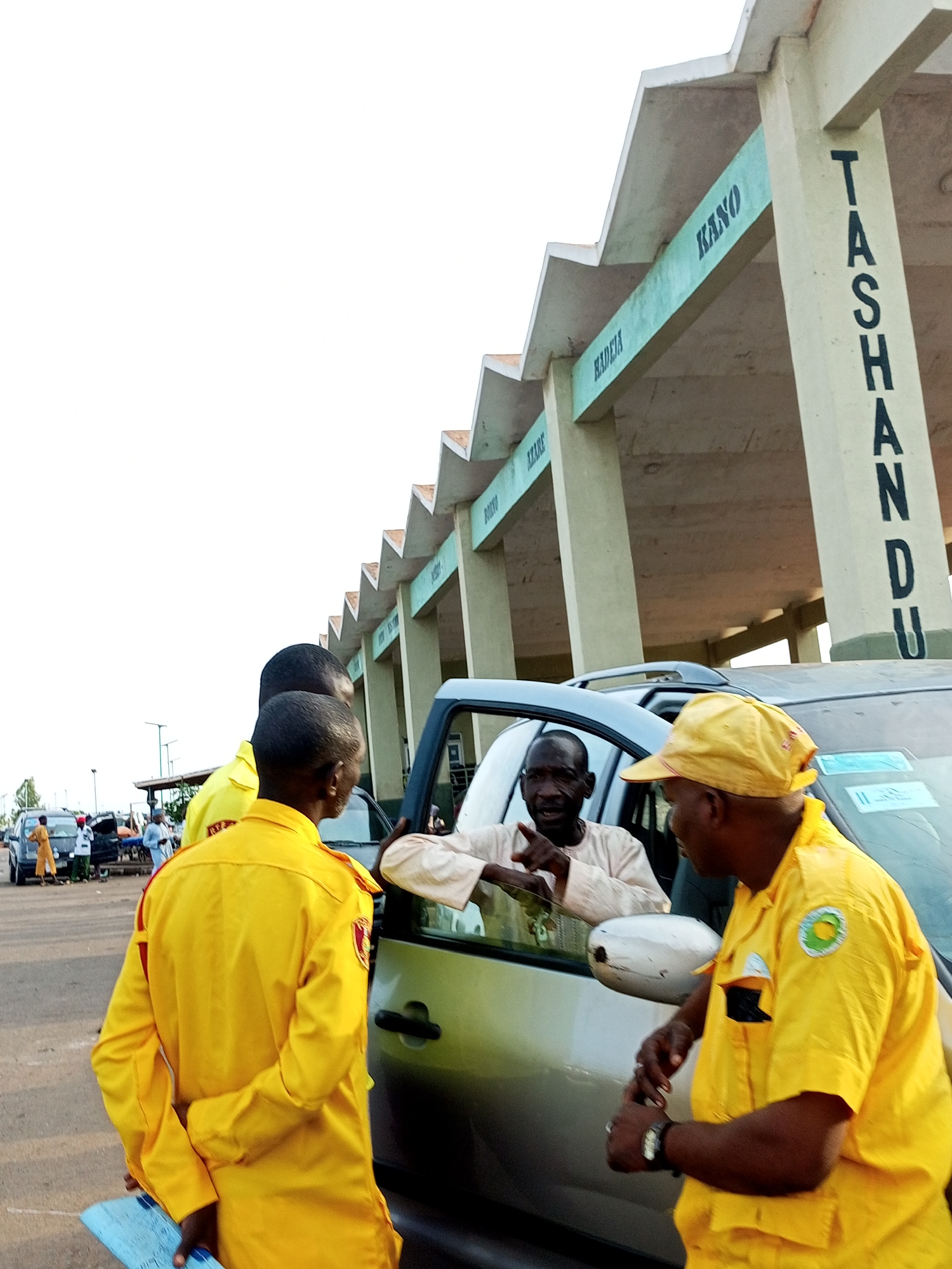 A group of men in yellow uniforms are gathered around a car, suggesting a potential interaction or transaction. One man in the driver's seat appears to be speaking, gesturing with his hand. Another man in a yellow uniform, facing away from the camera, is holding a blue document. The environment appears to be an outdoor waiting area, possibly a bus or taxi park, with a building featuring pillars with the names of cities like "Kano" and "Tashandu" painted on them. This architectural detail strongly indicates the location is likely within Gombe, Nigeria, given the prevalence of such signage in transportation hubs in that region, which often denote routes or destinations. Several other vehicles and people are visible in the background, hinting at a busy public space. The sky is overcast, and the lighting suggests it is daytime.