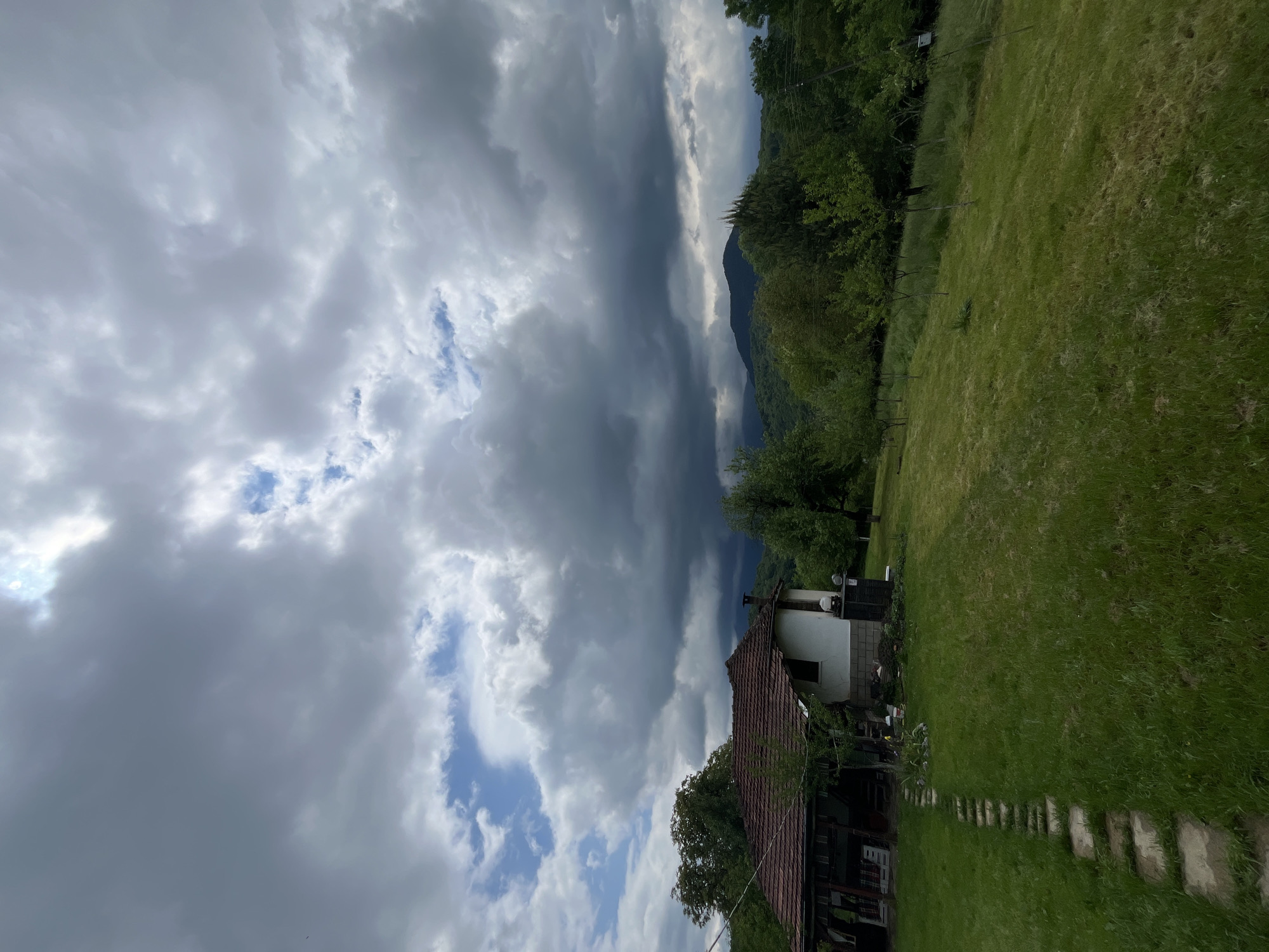 The image shows a rural scene in Shopite, Bulgaria. A weathered house with a reddish-brown tiled roof sits in a grassy field. A stone pathway leads away from the house. Behind the house, a dark green wooded area rises towards a distant mountain. The sky is filled with heavy, grey storm clouds, though patches of blue sky are visible. The photograph appears to be taken at an angle, possibly to accommodate the cloud formation. No people are visible.