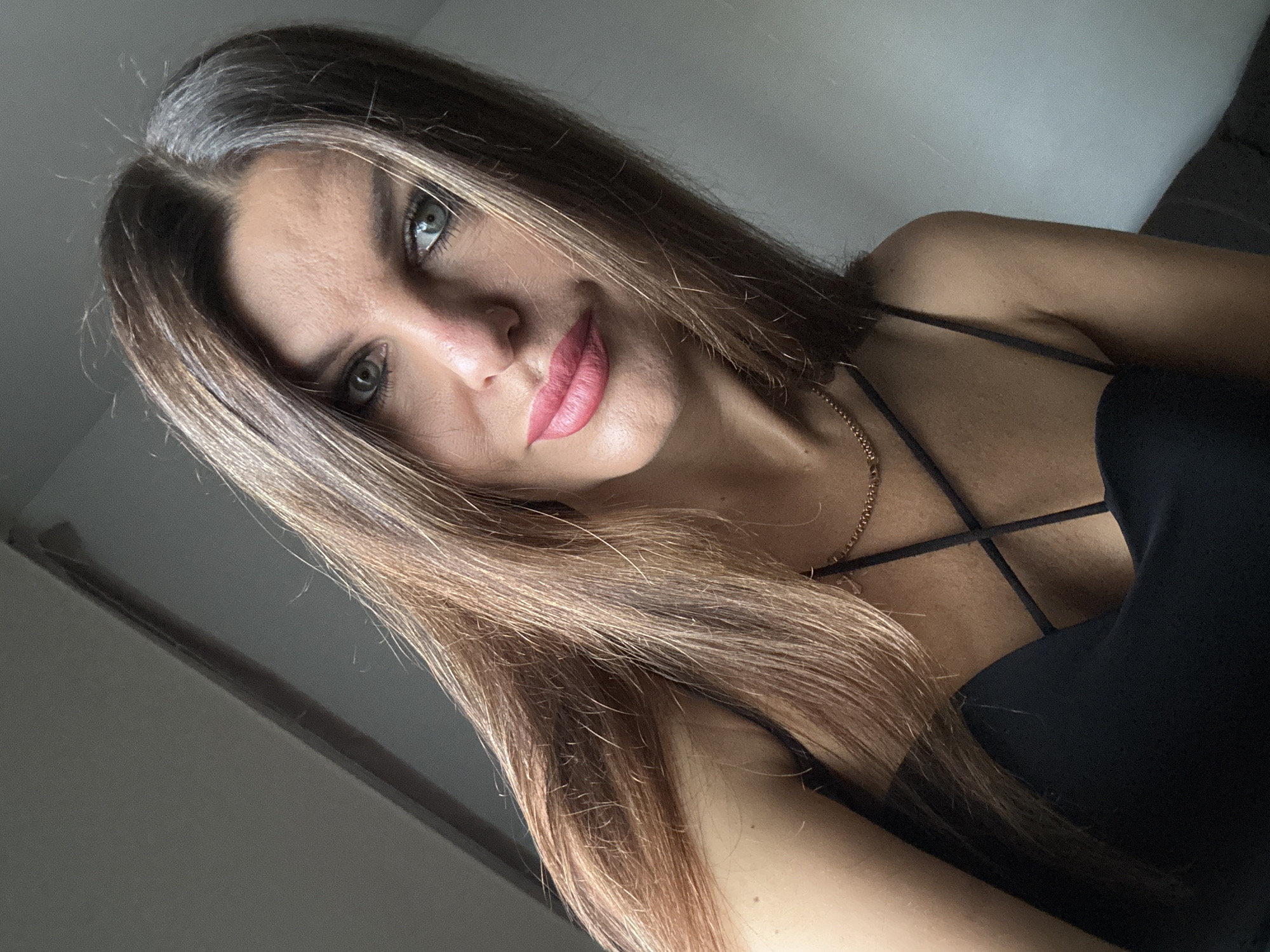 A medium shot selfie of a woman with long, layered brown hair and striking green eyes. She is looking directly at the camera with a slight smile and pink lipstick. She is wearing a black spaghetti strap top and a delicate gold chain necklace. The background is dimly lit with a grey wall and a hint of natural light coming from the left. There is no indication of the city of Chaikovsky, Russia in this image.