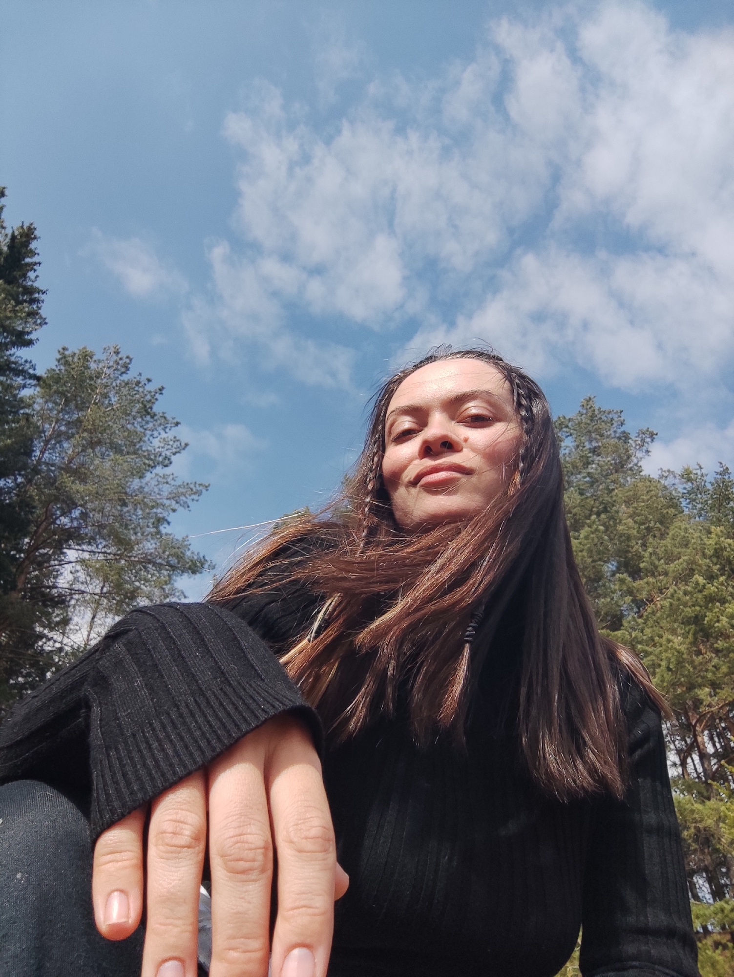 A medium shot captures a woman with long, dark brown hair, a small mole on her right cheek, and several small braids framing her face, gazing downwards at the camera with a slight smile. She is wearing a black ribbed sweater. Her left hand is partially visible in the foreground.
The background includes pine trees, partially visible in the lower left and right corners of the frame, suggesting an outdoor location. Above the trees, a blue sky is visible with scattered white clouds. The perspective suggests that the photo was taken from a low angle, looking upwards.
The location is inferred to be Фоки, Russia based on the user's specification. No specific landmarks or details within the image confirm the location.
