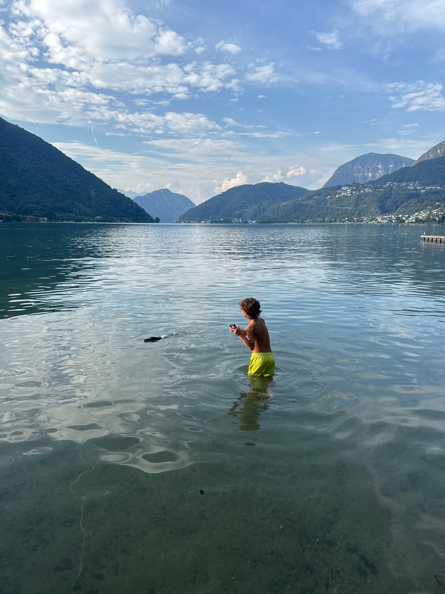 A lone young boy, wearing yellow swim trunks, stands in the shallow water of a calm lake. He is looking out at the water, holding a dark object in his hands. In the distance, a small, dark, remote-controlled boat is visible on the water, leaving a small wake. The lake is surrounded by lush green mountains, with some buildings dotted along their slopes. The sky is a clear blue, with scattered white clouds. The overall scene is peaceful and serene, suggesting a warm summer day. The location is likely Lake Lugano in Ticino, Switzerland, a region known for its beautiful lakes and mountainous scenery.