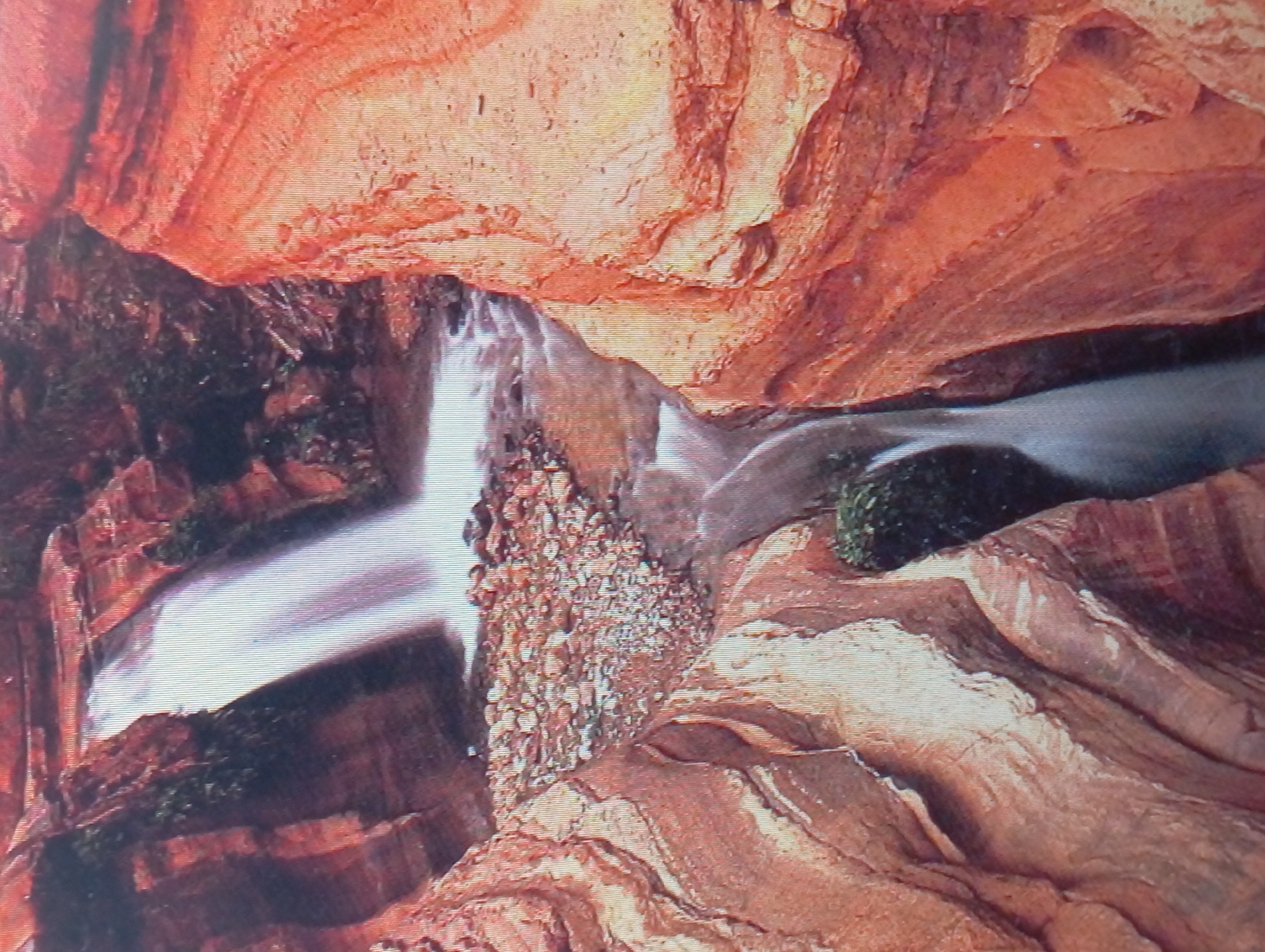 The image depicts a slot canyon with a waterfall and stream. The canyon walls are layered reddish-brown rock formations. Water cascades down the waterfall into a bed of rocks and then flows through the canyon. Patches of vegetation are growing on the canyon walls. The location is not in Karaganda, Kazakhstan.