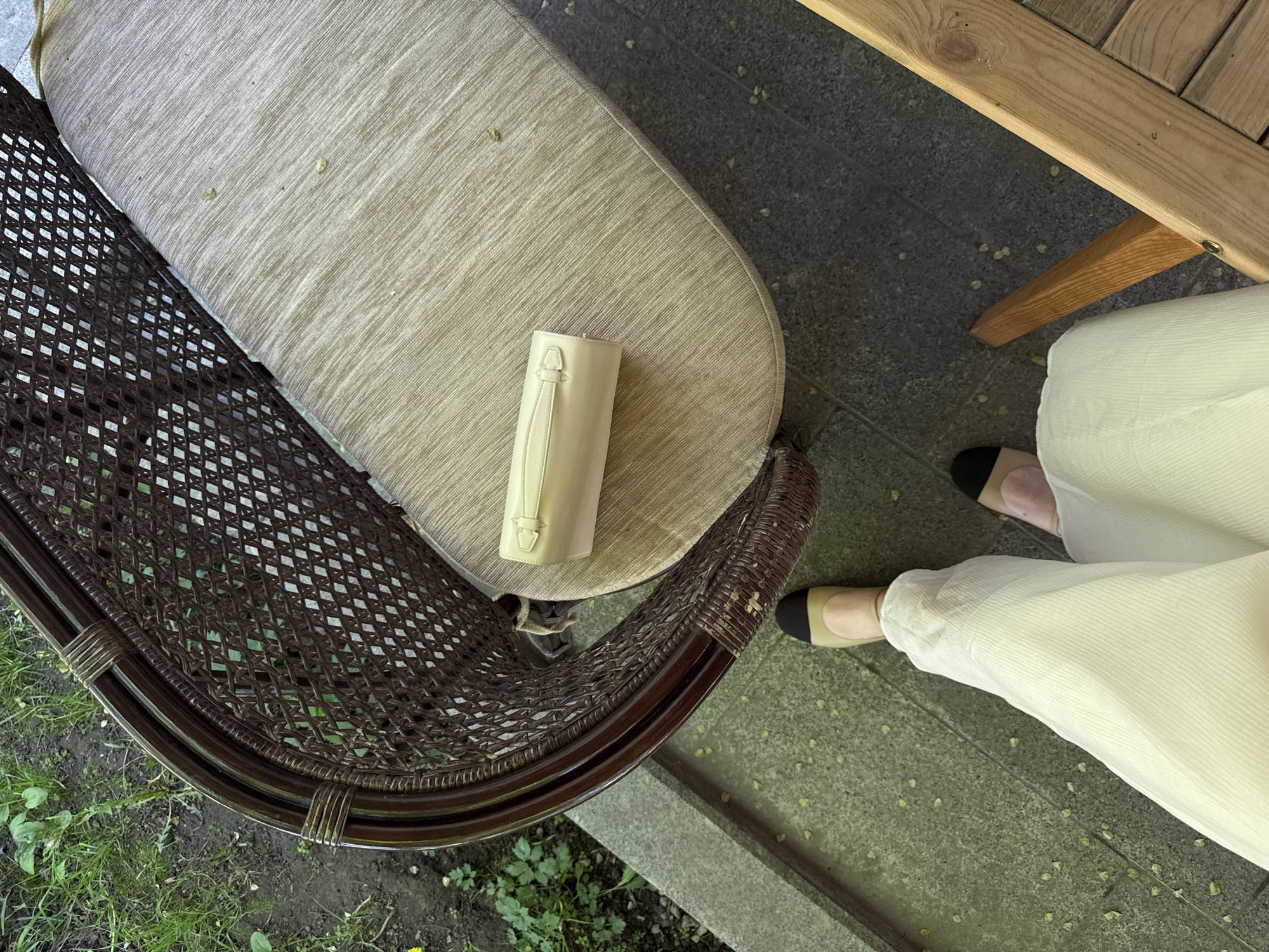 The image depicts a partially visible outdoor scene, including a portion of a wicker chair with a light brown cushion, a light-colored cylindrical object with a handle on the cushion, part of a wooden table, and a person's legs wearing white pants and two-tone flats. The setting appears to be a patio or garden area with concrete slabs and some vegetation visible near the chair. The exact location within Moscow, Russia, cannot be determined from the provided image.