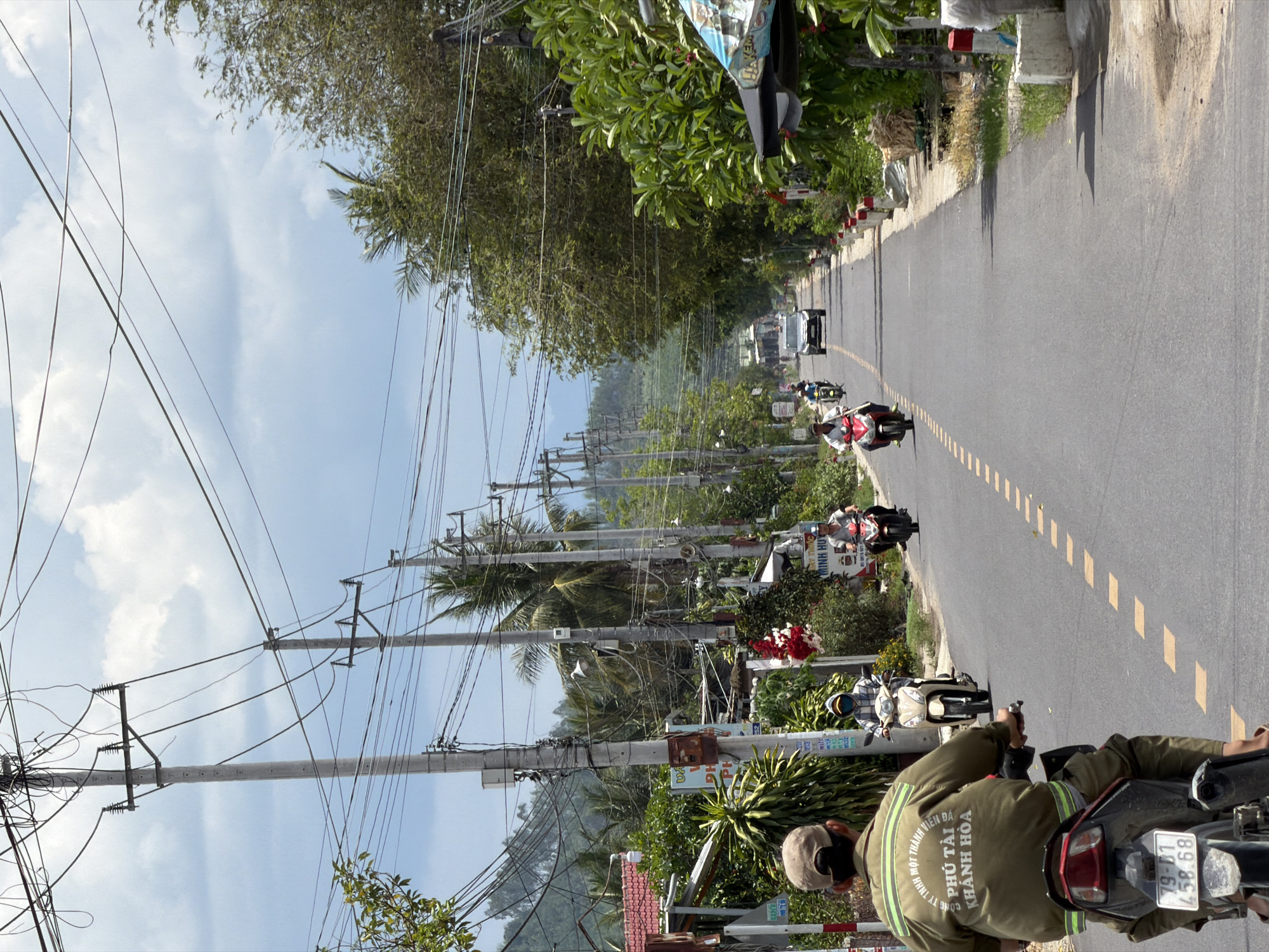 The image depicts a street scene in Diên Thọ, Vietnam, under a bright, overcast sky. In the foreground, a person wearing a tan cap and a khaki jacket is riding a motorcycle. The motorcycle has a license plate with the number "79-D1 458.68". The jacket has text printed on the back in Vietnamese.
The road ahead is paved and lined with dashed yellow markings. Several other individuals are riding motorcycles in the same direction. A dark-colored SUV is visible further down the road, also heading away from the viewer.
The sides of the road are lined with numerous utility poles carrying a complex network of electrical wires. Lush green vegetation, including trees and bushes, grows on both sides of the road. Some buildings with red tile roofs are partially visible through the foliage. Red and white markers are placed along the edge of the road. The overall impression is of a moderately busy rural or suburban street in Vietnam.