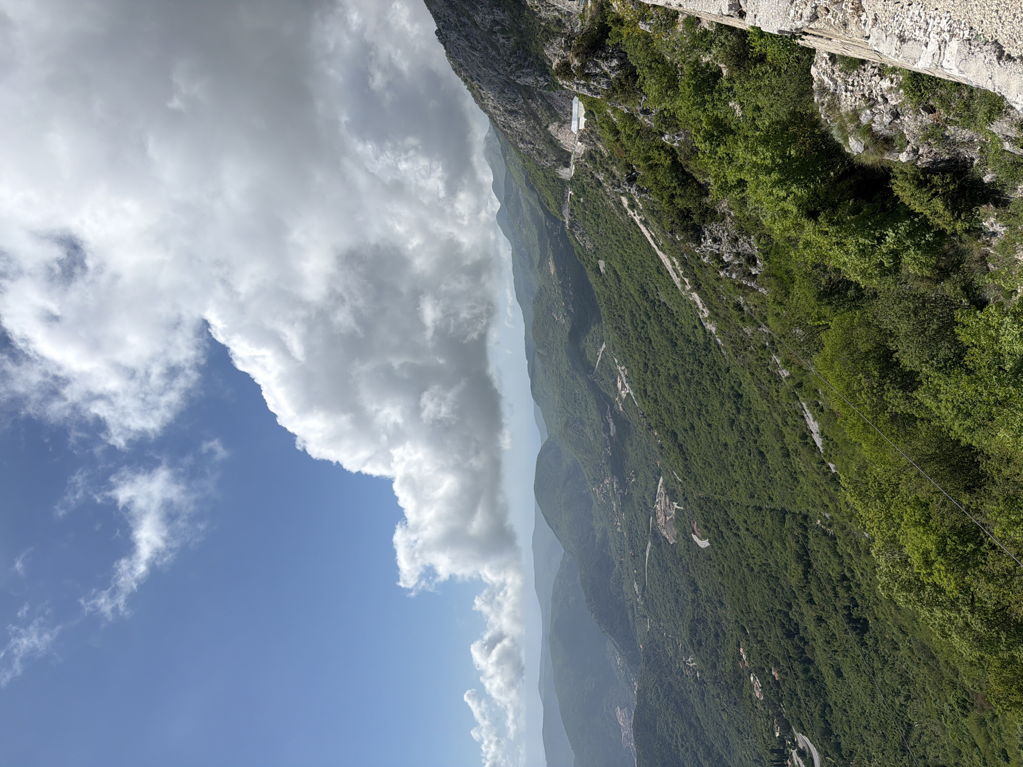 The image depicts a mountainous landscape in Brajici, Montenegro. A large cloud formation dominates the left side of the frame, juxtaposed against a blue sky. The mountains are covered in dense green vegetation. A stone structure or wall is visible at the top right corner. The scene appears to be taken from a high vantage point.