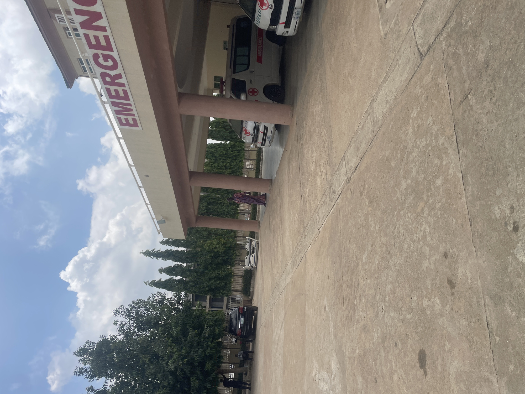 The image shows the exterior of an emergency department at a hospital in Abuja, Nigeria. A large, red sign with the word "EMERGENCY" is visible on the building's facade. In front of the building, a covered entryway shelters two white ambulances, one with "AMBULANCE" written on its side. To the left, a black SUV is parked, and several people are standing or sitting nearby. A woman in a patterned dress walks in the foreground. The ground is paved with concrete, and trees line the background. The sky is blue with scattered clouds.
