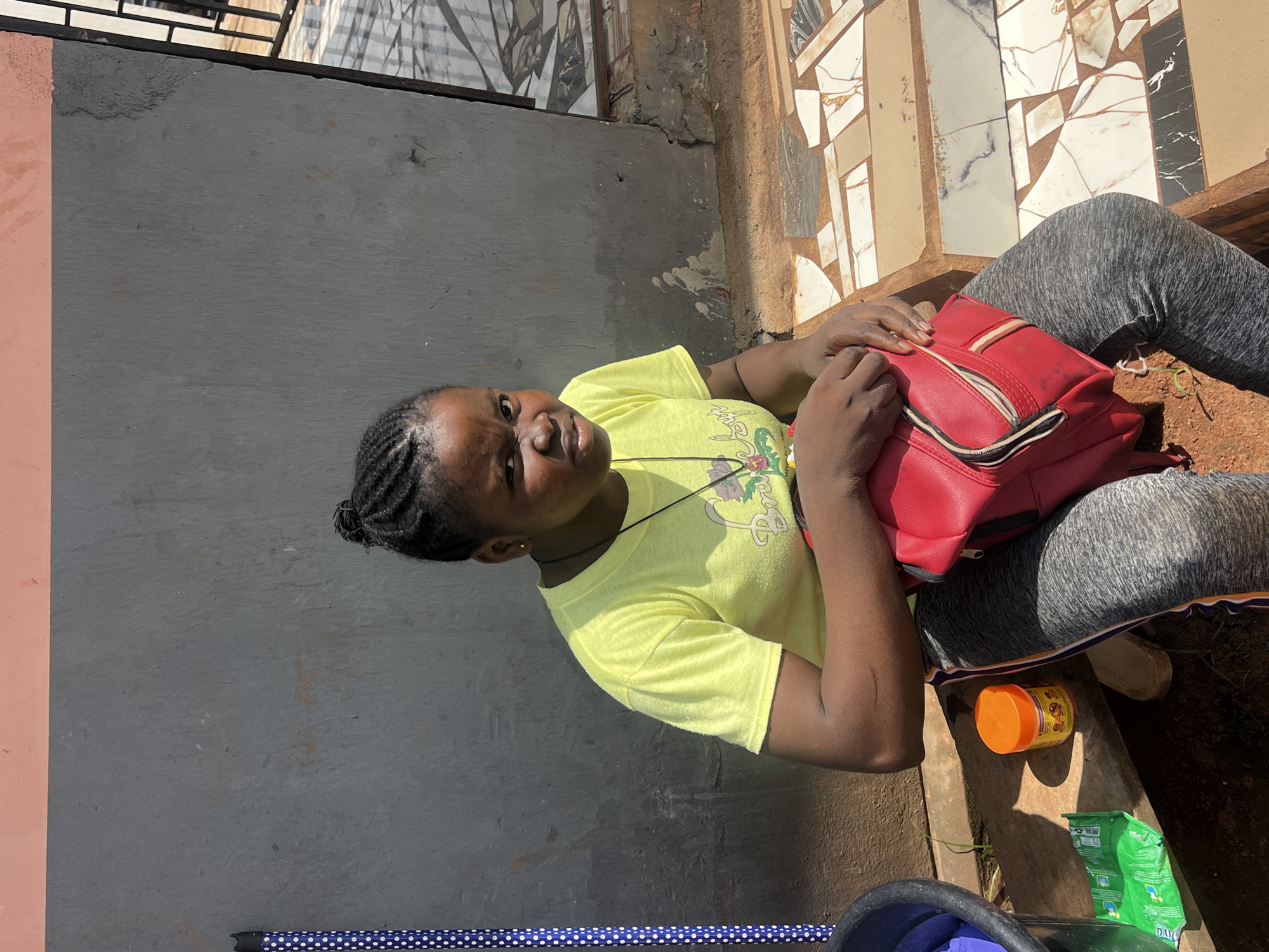 In Nkpor, Nigeria, a young woman is pictured sitting on a step outdoors. She is wearing a bright yellow t-shirt and grey leggings with a white stripe down the side. Her hair is styled in braids tied back. She holds a red backpack on her lap with both hands. Beside her on the step sits an orange container and a green packet. A blue and white polka-dotted pole is visible on the left edge of the image, and a black bucket is partially visible. The background consists of a grey wall with a pink stripe at the top, and a tiled pathway to the right. The ground is a mix of dirt and small stones.