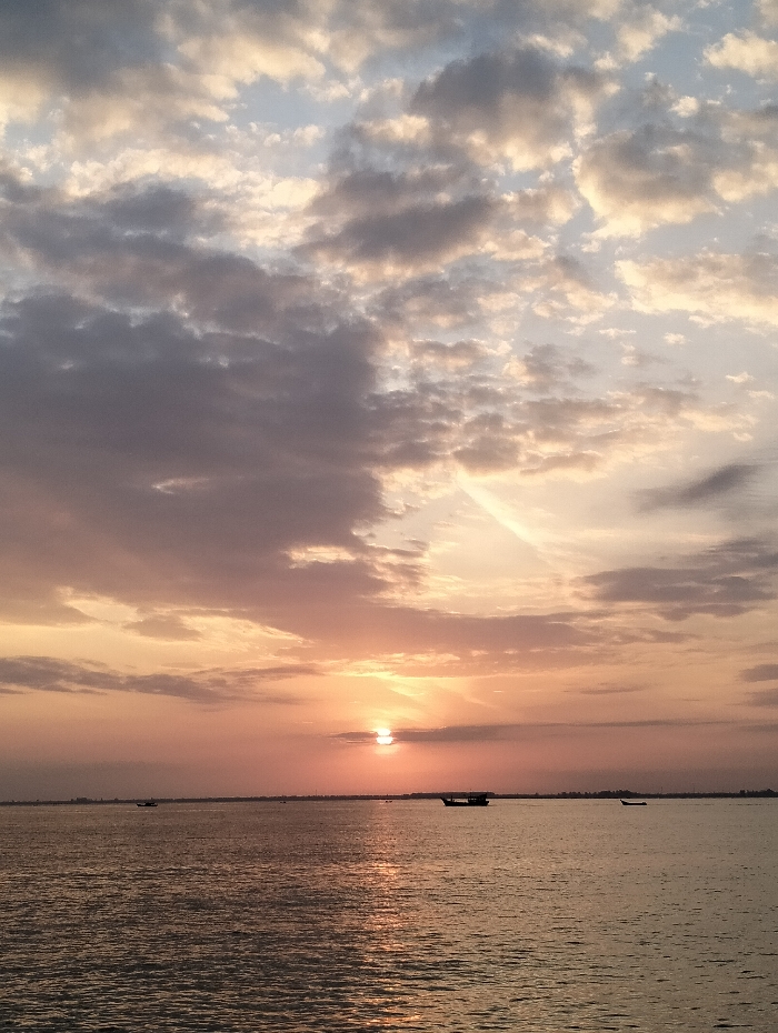 A serene sunset over a tranquil body of water. The sky is filled with clouds tinged with shades of orange, pink, and gray, while the sun is peeking through the horizon line. Three boats are scattered across the water, their dark silhouettes contrasting with the luminous reflections dancing on the water's surface. The scene evokes a sense of peace and quiet beauty.