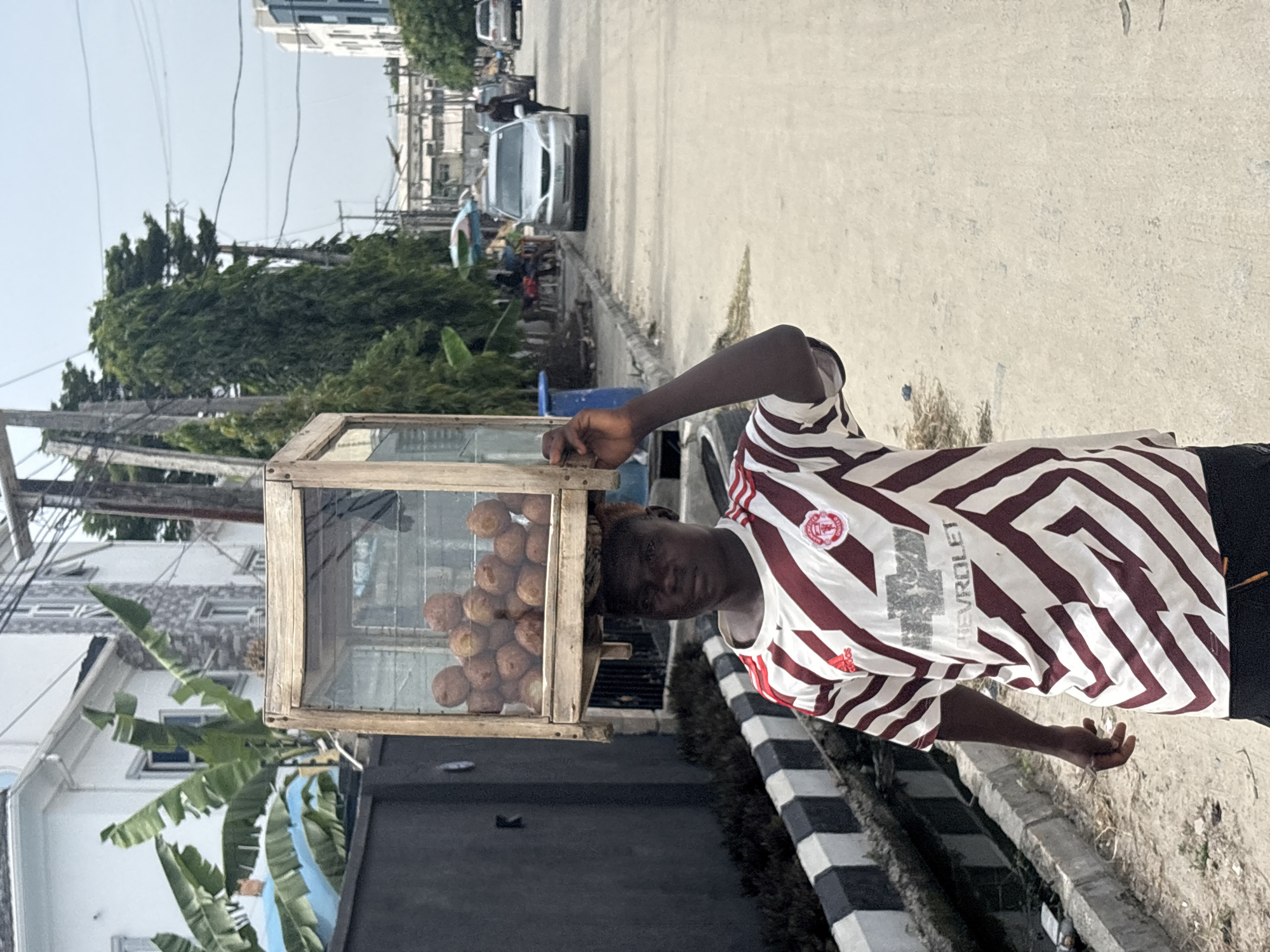 An individual, appearing to be a young adult male, is depicted walking along an unpaved road in Igboefon, Nigeria, under an overcast sky. He wears a white and maroon/red striped short-sleeve shirt with an Adidas logo, Manchester United crest, and "Chevrolet" text. On his head, he carries a rectangular wooden display box with glass sides, filled with numerous round, brown food items, likely fried dough. His left hand is raised, lightly touching the box for balance. To his left, a black and white striped concrete curb borders a narrow drainage ditch, behind which is a dark grey wall and a multi-story building with visible green foliage. The background reveals additional residential or commercial buildings, utility poles with overhead wires, tall trees, and several parked or moving vehicles on the road, with indistinct figures further down.