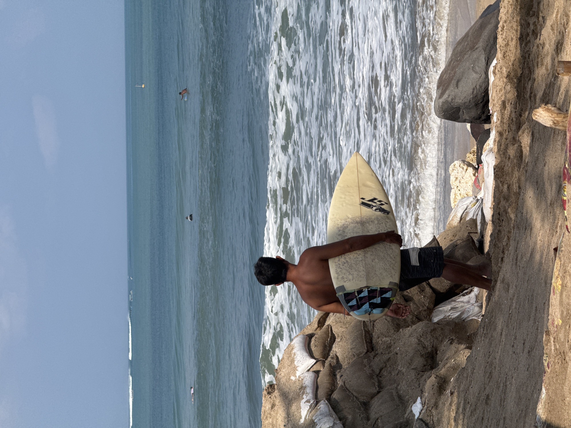 A man stands on a sandy beach, facing the ocean, holding a surfboard. He is shirtless, wearing dark patterned swim trunks. The surfboard is light-colored with some colorful abstract designs on the tail. Behind him, a steep embankment is reinforced with numerous sandbags. The ocean stretches out to the horizon under a clear blue sky. Several surfers are visible in the water, some paddling and one possibly on a wave in the distance. The waves are breaking near the shore, creating white foam. The lighting suggests it is daytime. The scene depicts a typical surfing location in Kuta, Bali, Indonesia.