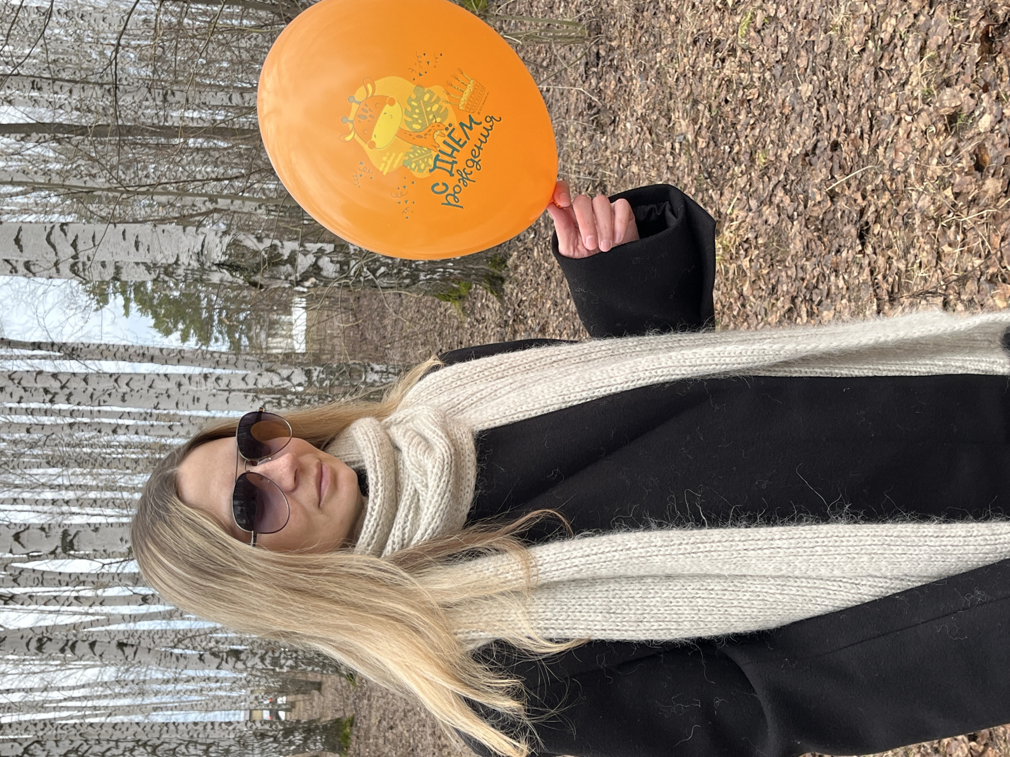A woman with blonde hair and sunglasses is standing in what appears to be a park in Chaikovsky, Russia, holding an orange balloon with a drawing and inscription on it. The background shows a birch tree forest and the ground is covered with fallen leaves. She is wearing a black coat and a white scarf. The picture is taken outdoors, likely in daylight.