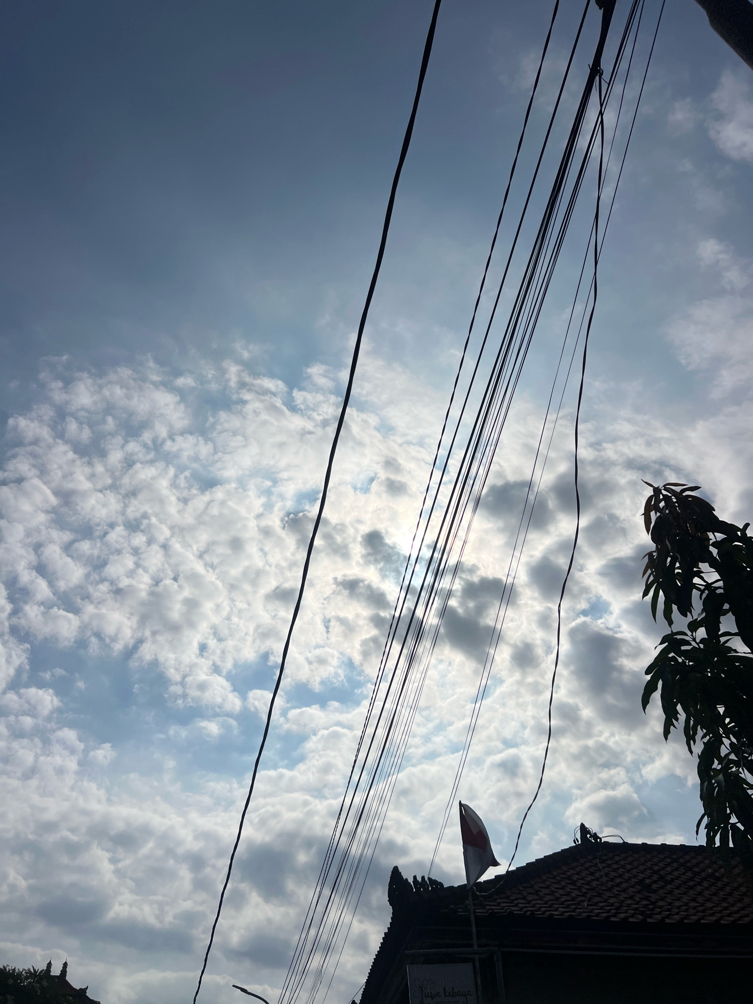 The image captures a view of the sky from Kuta, Indonesia, characterized by a dramatic interplay of light and shadow. A dense cluster of thin, dark electrical wires crisscrosses the frame, obscuring parts of the bright, cloudy sky. The sun is partially visible behind the clouds, casting a gentle glow. In the foreground, the tiled roof of a building is silhouetted against the sky, topped with an Indonesian flag, its red and white colors distinct against the muted background. To the right, the dark silhouette of tree foliage adds another layer to the scene. The overall impression is one of a typical urban landscape in Kuta, with the natural elements of sky and sun juxtaposed with the man-made structures of wires and buildings.
