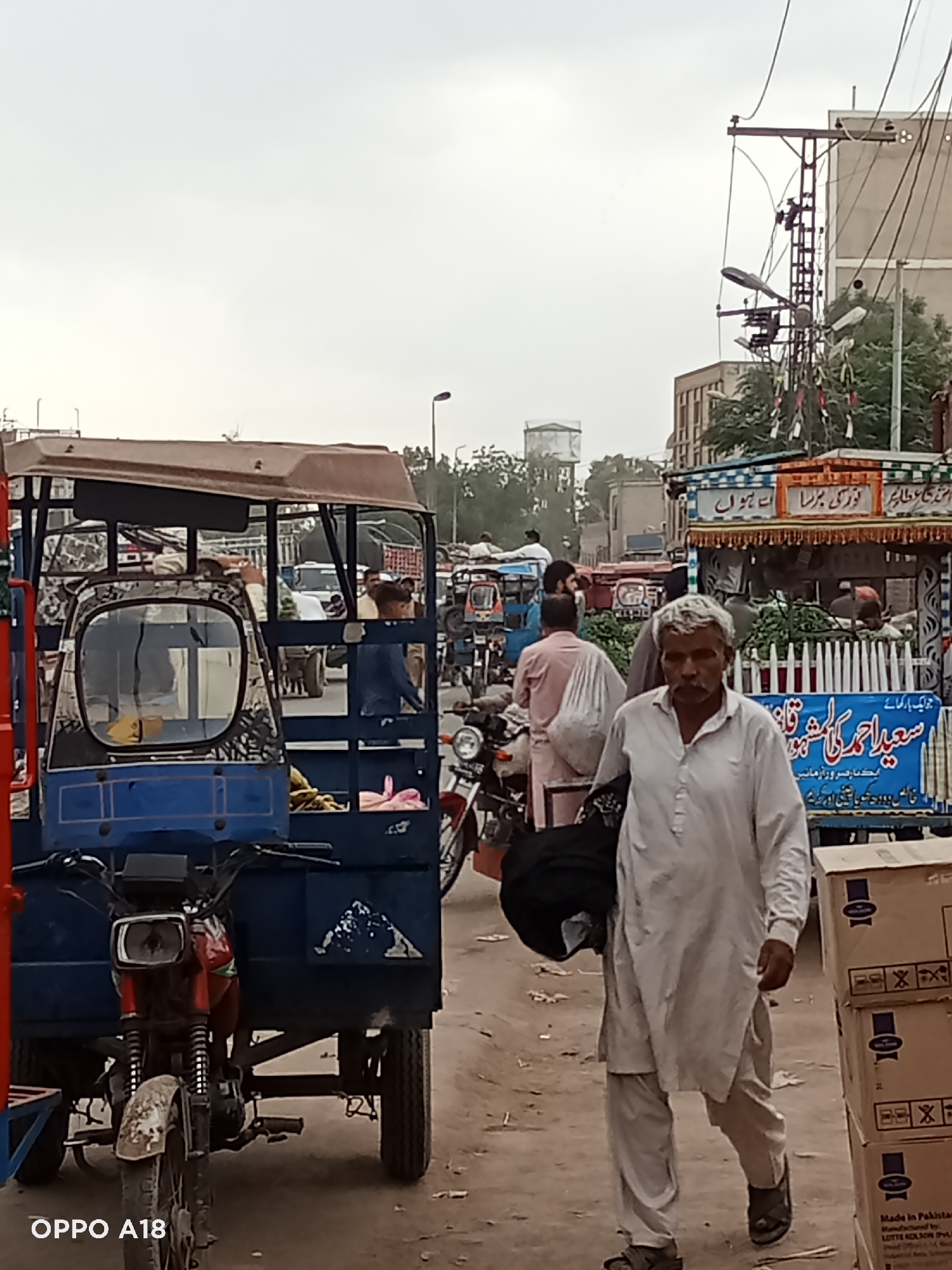 The image depicts a street scene in an urban environment, possibly a marketplace. A man in traditional Pakistani attire (white shalwar kameez) is walking towards the camera. Several auto-rickshaws and motorcycles are present, suggesting a busy and congested area. A vendor stall with vegetables is also visible. The sky appears overcast. Text in Urdu is visible on signage, implying the location is likely in Pakistan.