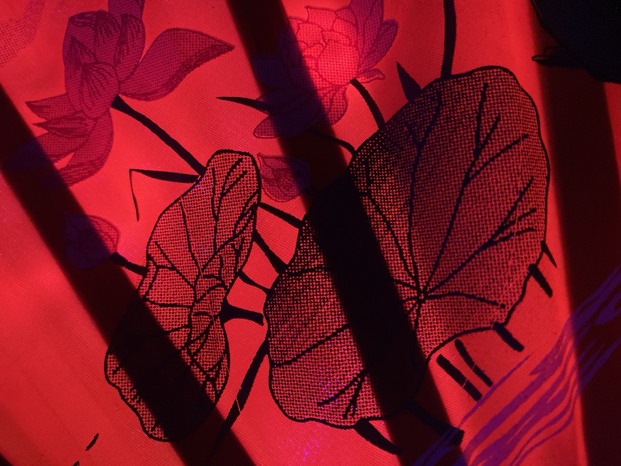 A close-up shot depicts a fabric with floral and leaf patterns in shades of red and black. The patterns resemble lotus flowers and leaves, stylized in a stippled, halftone-like texture. Light and shadow play across the fabric, creating sharp contrasts. Strong, dark lines cut diagonally across the image, suggestive of an interior setting. The overall mood is somewhat dramatic due to the bold colors and strong shadows. Given the prompt, the location is likely somewhere in Baan Kai, Thailand.