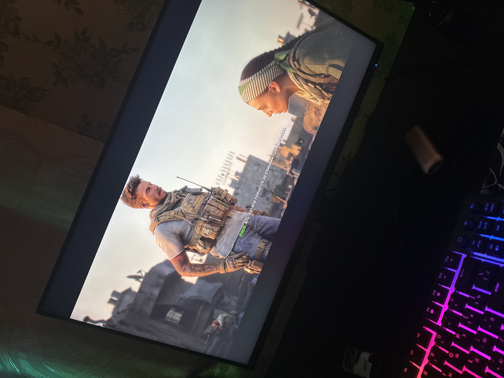 The image shows a TV screen displaying a scene from a video game, possibly Call of Duty: Modern Warfare. Two characters are visible: a male soldier in tactical gear and a female soldier with a headscarf. The background appears to be a war-torn city, with damaged buildings. The TV screen is tilted, and part of a gaming keyboard with RGB lighting is visible in the foreground. The scene is likely set in a fictional Middle Eastern or North African location, not Makiivka, Ukraine.