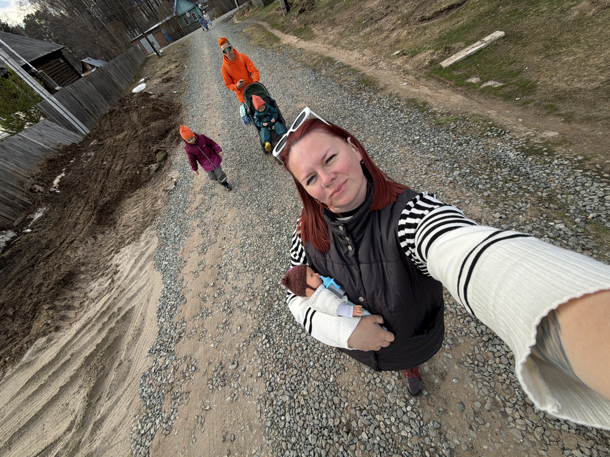 A high-angle, wide-angle selfie captures a group of people, likely a family, walking on a gravel and dirt road in Chaikovsky, Russia. In the foreground, a woman with auburn hair and a nose ring, wearing a black vest over a white and black striped long-sleeve shirt, takes the photo. White sunglasses rest on her head, and she holds a baby doll wearing a knitted brown hat and holding a blue bottle.
Behind her, a man in a bright orange hoodie and sunglasses pushes a stroller containing a child dressed in green with an orange hat. Alongside the stroller, another child in a purple jacket and orange hat walks. Further down the road, another person is visible in the distance.
The setting appears to be a rural or semi-rural area, characterized by unpaved roads. To the left, a wooden fence runs alongside a large mound of dark, moist soil and sand, with some tire tracks visible. The right side of the road features a grassy bank with a concrete slab. Wooden houses and bare trees are visible in the background, suggesting early spring or late autumn. The weather seems cool, as everyone is dressed in warm attire. The woman in the foreground has a slight smile, indicating a content mood during what appears to be a casual family outing. No explicit text is visible in the image.