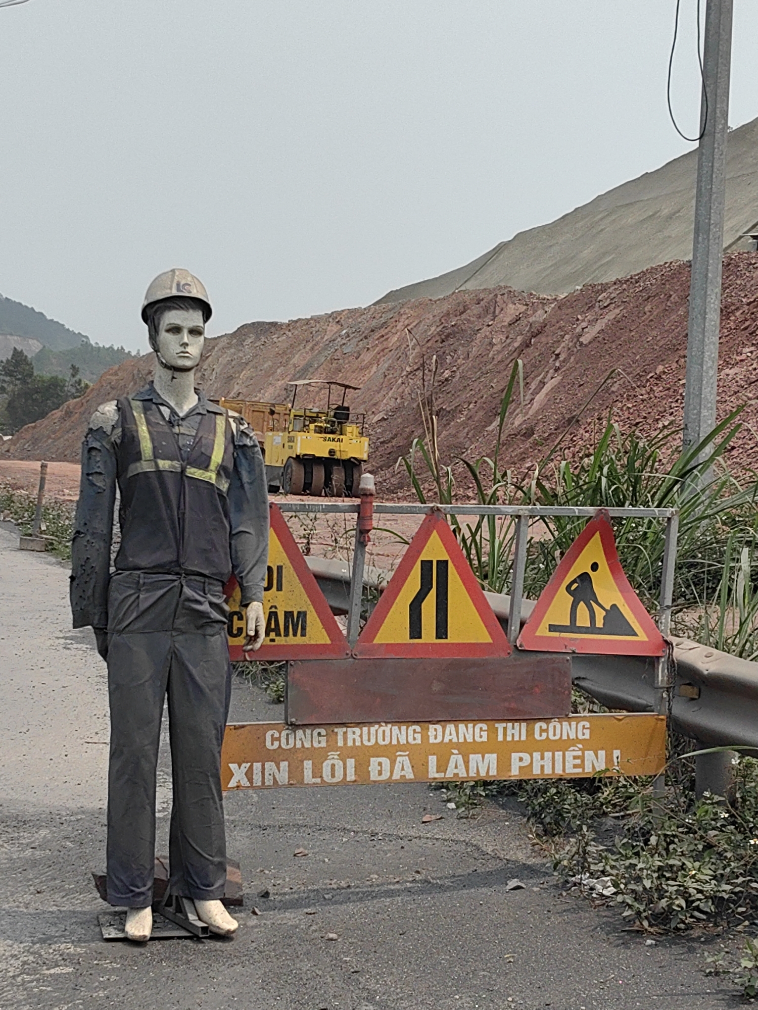 A construction site in Vũ Oai, Vietnam, is depicted. A mannequin in a worn construction worker's uniform and hard hat stands to the left, positioned in front of construction signs. These signs, painted yellow and red, include warnings about road construction and reduced lanes. Behind the mannequin and signs, a large dirt hill or embankment is visible, along with construction equipment. The road surface is asphalt. Vegetation is sparsely present along the road's edge. A utility pole stands further back.