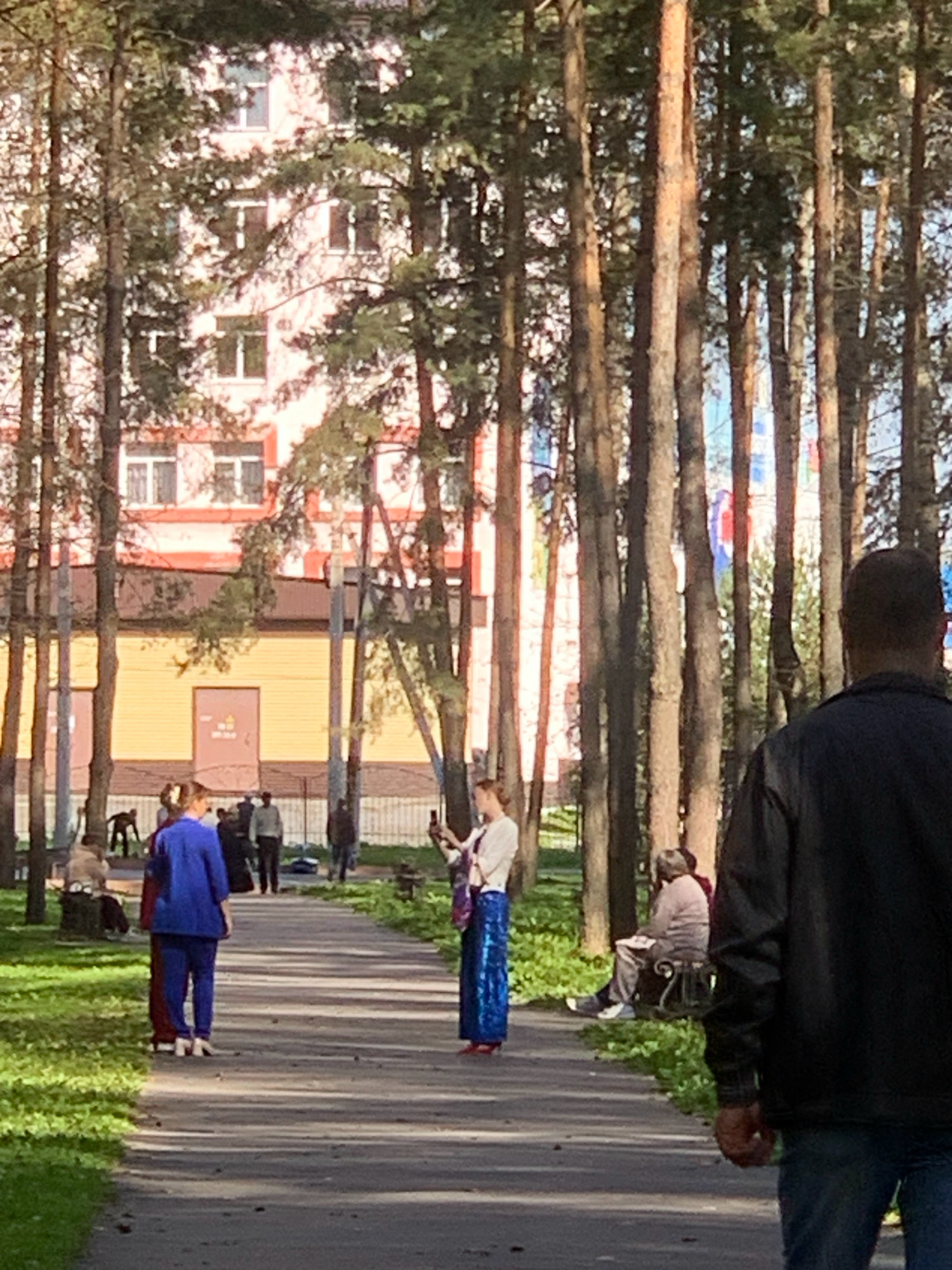 The image depicts a park-like setting with tall trees lining a paved walkway. In the background, a multi-story building with a pink facade and numerous windows is visible. The building appears to be an apartment complex or similar residential structure.
Several people are present in the scene. On the left side of the walkway, a woman in a blue outfit is standing, facing away from the camera, with another person in a dark red garment beside her. Further back, several individuals are seated on benches or standing, engaged in various activities.
In the center of the image, a woman is standing and taking a photo with her phone. She is wearing a white top and a long, shimmering blue skirt. To the right, a person is seated on a bench. In the foreground, the back of a person in a black jacket and blue jeans is visible, partially obscuring the view.
The environment appears to be a park or a public garden, with green grass and foliage interspersed with trees. The lighting suggests it is daytime, possibly late afternoon, with sunlight filtering through the trees and casting shadows on the walkway. There are no specific identifiable landmarks that would confirm the location as Prokopyevsk, Russia.