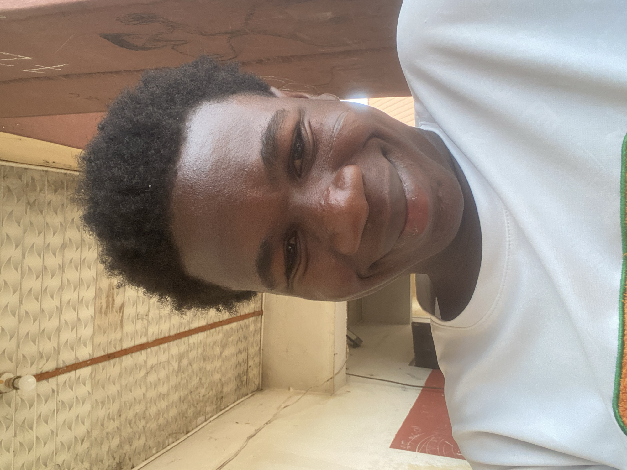 A dark-skinned young man with short, dark curly hair is smiling at the camera. He is wearing a white t-shirt with a green and orange design along the collar. He appears to be standing inside a building. The background shows a textured wall, white tiled flooring, and a glimpse of another room with a dark object. The location is Tudun Wada Bibinu, Nigeria.