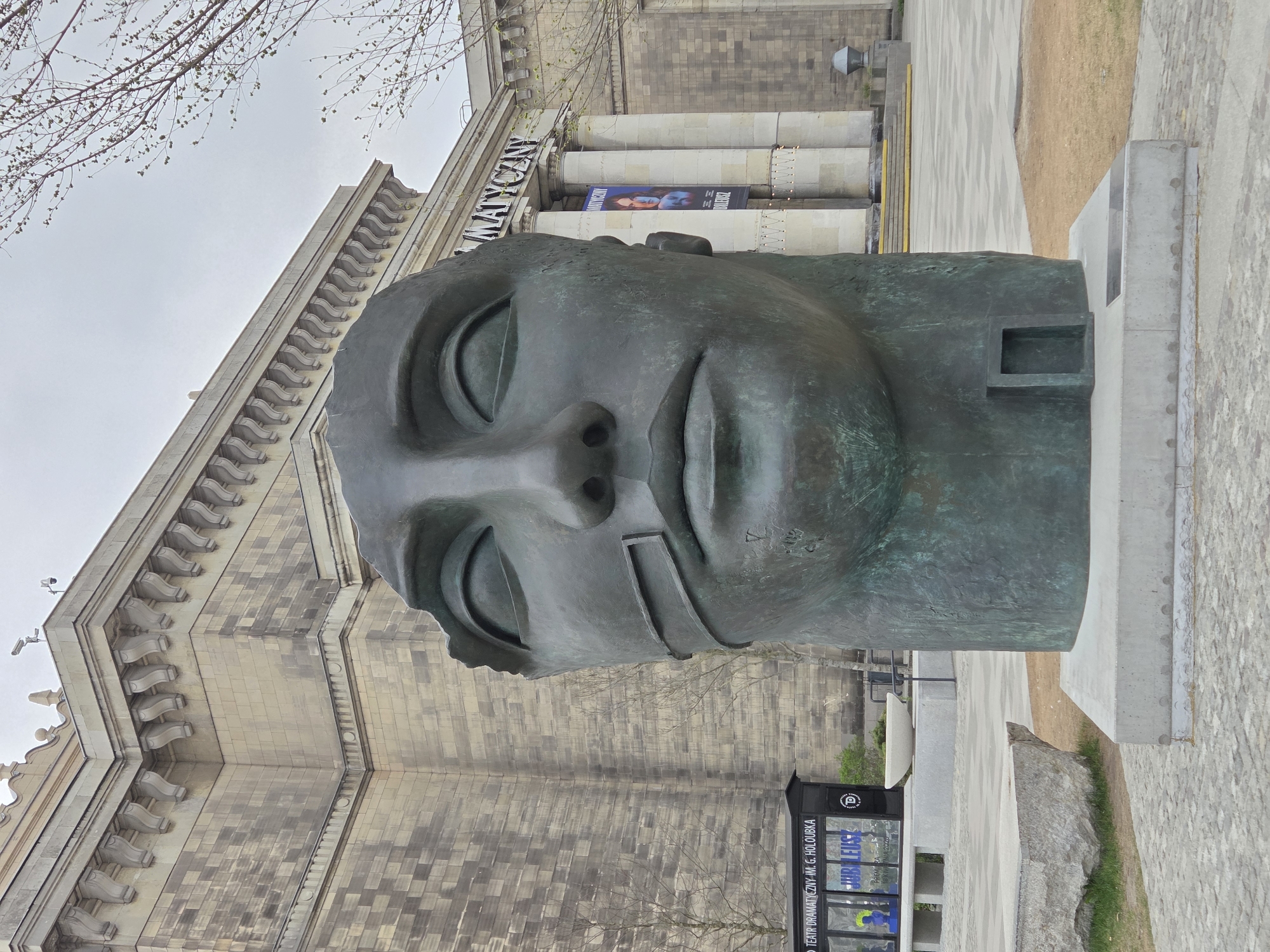 The image shows a large, bronze head sculpture in front of the Palace of Culture and Science in Warsaw, Poland. The head is a stylized representation of a face with closed eyes, a prominent nose, and a rectangular cut-out on the left side of the face. The sculpture is mounted on a concrete base. Behind the sculpture is the Palace of Culture and Science, a large, imposing building with a detailed facade. A banner is displayed across the front of the building. To the side of the sculpture is a pedestrian walkway and some greenery. A small building or structure with text in Polish is visible on the right side of the frame. The sky is overcast.