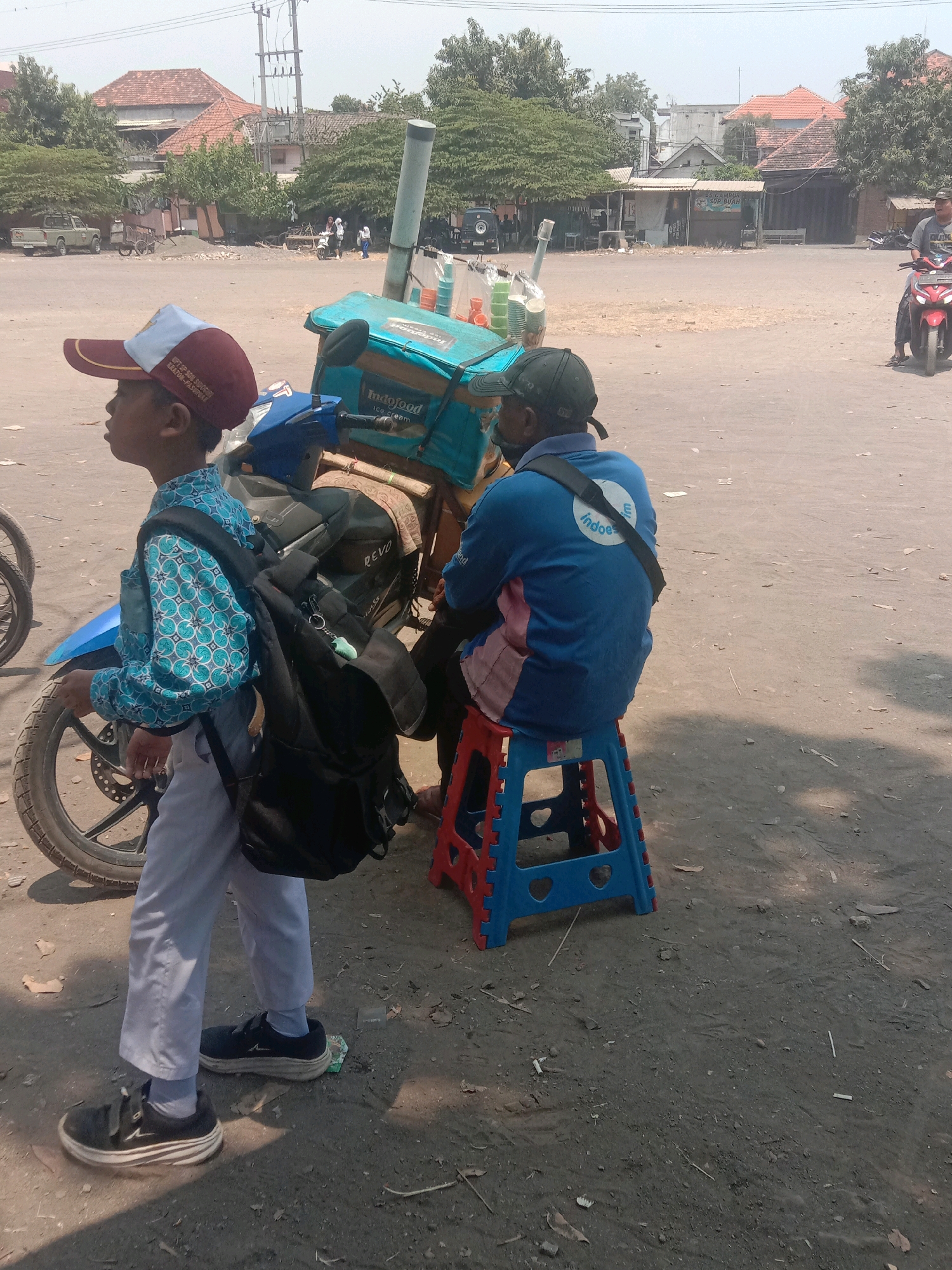 In the city of Ngempit, Indonesia, an outdoor scene depicts a dusty, sun-drenched area with several individuals and a motorcycle. In the foreground, a young boy, wearing a blue patterned shirt, white pants, a red and white cap, and a black backpack, stands near the rear wheel of a blue motorcycle. The motorcycle is equipped with a turquoise delivery box labeled "Indofood Ice Cream." To the right of the boy, a man in a blue long-sleeved shirt with "Indofood" printed on the back is seated on a red and blue plastic stool. He wears a dark baseball cap and a black bag strap across his chest. In the mid-ground, a red motorcycle is ridden by an individual wearing a dark helmet, heading away from the camera. The background reveals a street lined with buildings, trees, and utility poles. The ground is covered in dirt and scattered debris.