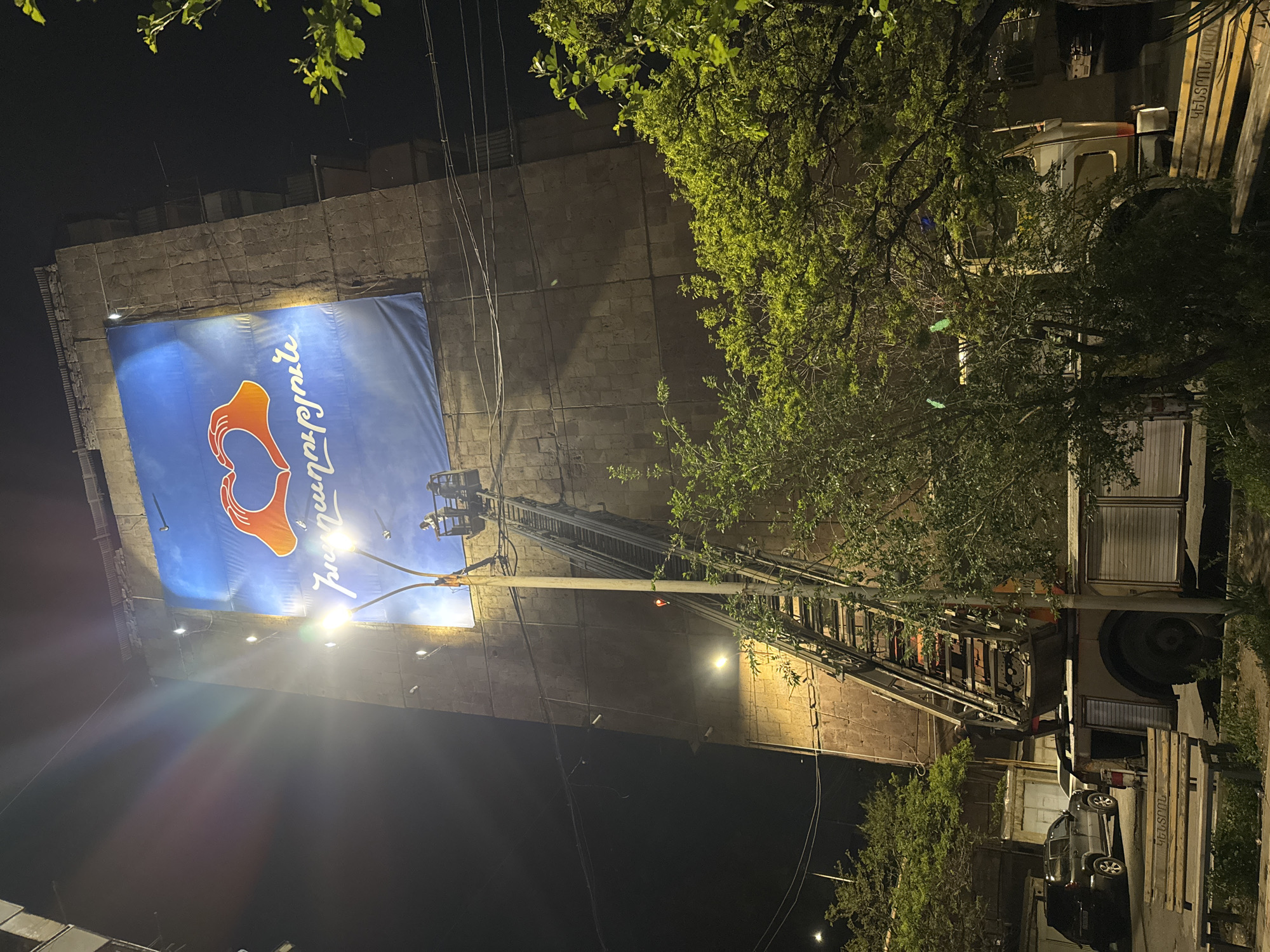 At night, a tall, multi-story building made of rough-hewn stone is illuminated by several light sources as workers are on a platform at the end of a tall ladder, working on a large banner of a blue background with an orange logo of two hands cradling something. The banner also has white text in a foreign language. A fire truck is parked along the right side of the building, and its ladder is extended to the platform. A streetlight is shining from just beyond the fire truck, and tree branches frame the building. A few cars are parked on the street next to the building. The location is Yerevan, Armenia.