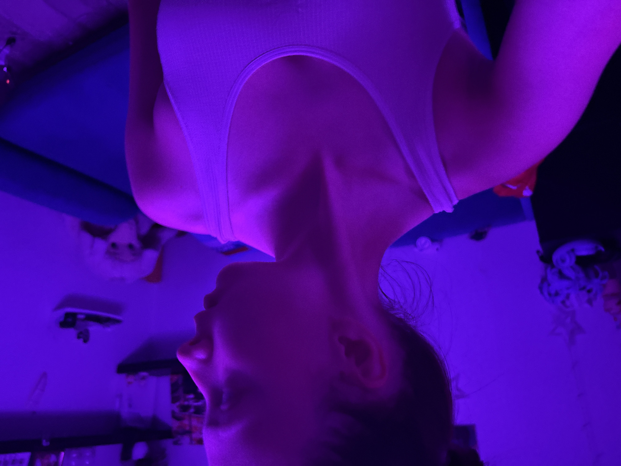 A woman wearing a white tank top is positioned in front of a room lit with purple lighting. The room includes a blue object, a counter, and various miscellaneous items. The location is identified as Прикамский, Russia. The image is oriented upside down.