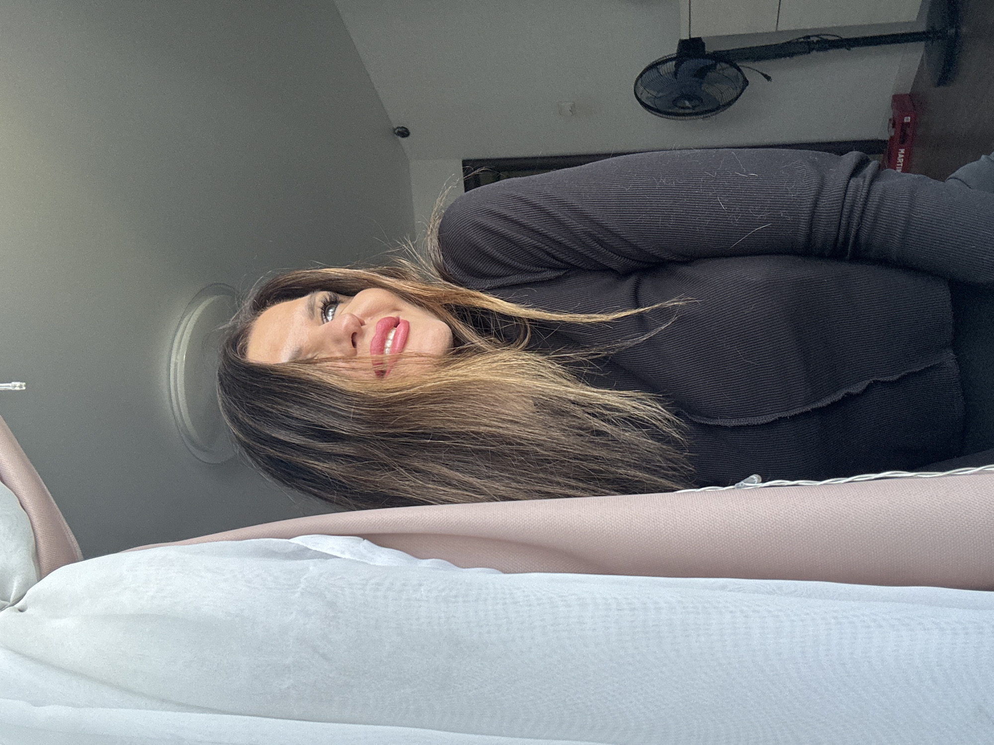 A woman with shoulder-length ombre hair is looking up and smiling. She is wearing a black, ribbed long-sleeved shirt. The foreground is blurry and shows a white and light pink fabric. Behind the woman, there is a ceiling with a circular molding. In the background, there is an indoor fan hanging on the wall and a red box. This image does not have sufficient data to specify the location in Chaikovsky, Russia.