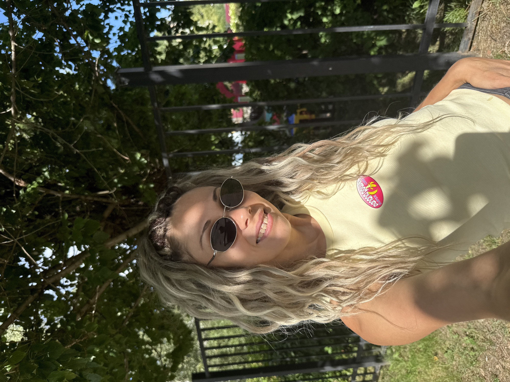 A blonde woman with wavy hair and sunglasses smiles at the camera. She is wearing a light yellow tank top with a pink and yellow oval patch on it that says "Banana Power." She is outdoors, with a black metal fence and trees behind her. The sun is shining, casting shadows on her arm. The location is the city of Rusanovo, Russia.