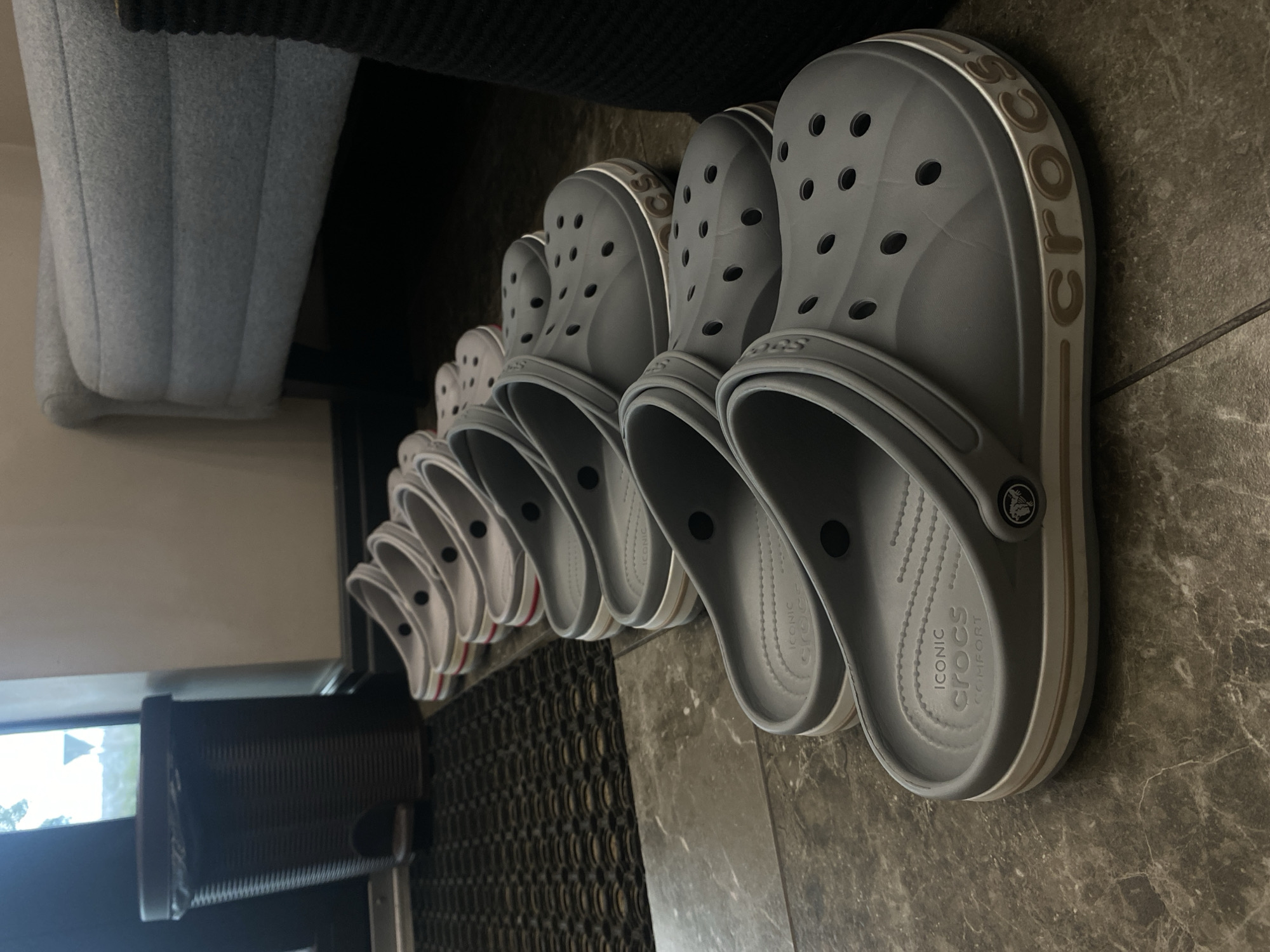 The image shows a line of grey Crocs shoes placed on a tiled floor. The shoes are arranged in a receding perspective, with the closest shoe being the largest and most detailed. The Crocs have the characteristic holes on the top and sides, and the "CROCS" logo is visible on the side of the shoe. Some of the shoes also have a white and brown trim. In the background, a grey upholstered chair and a black textured surface are partially visible. The setting appears to be indoors, possibly a hallway or entryway, given the floor tiling and the presence of what looks like a doormat. There are no people visible in the image, and the scene is static, with no activity taking place. The lighting suggests it is daytime, but no specific time of day can be determined. The location context provided is Votkinsk, Russia.