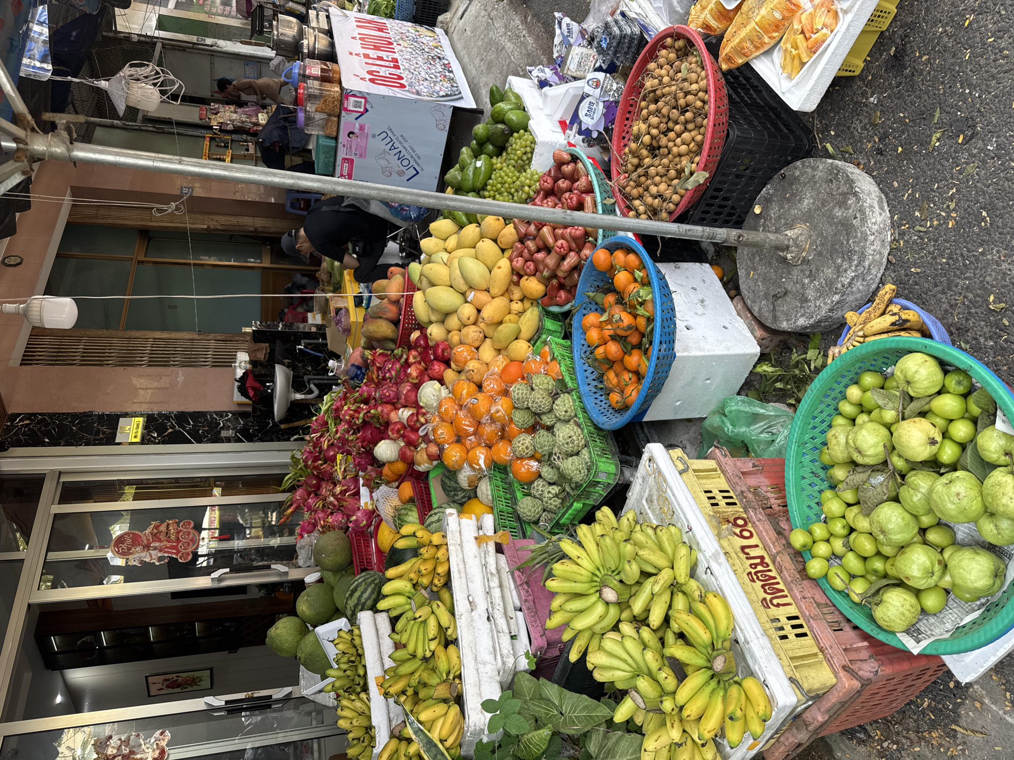 This image depicts a vibrant street-side fruit vendor in Đà Nẵng, Vietnam. A diverse array of fruits is displayed, including bananas, mangoes, oranges, dragon fruit, guavas, avocados, grapes, longans, and sugar apples. These fruits are arranged in baskets and crates, showcasing their colors and variety. A person is crouching near the fruits, likely attending to or arranging the produce. The backdrop includes the storefront of a building with glass doors, offering a glimpse into the interior. A sign with Vietnamese script is visible among the fruits. The setting is a typical urban street scene, characterized by a mixture of pedestrian and commercial activity.