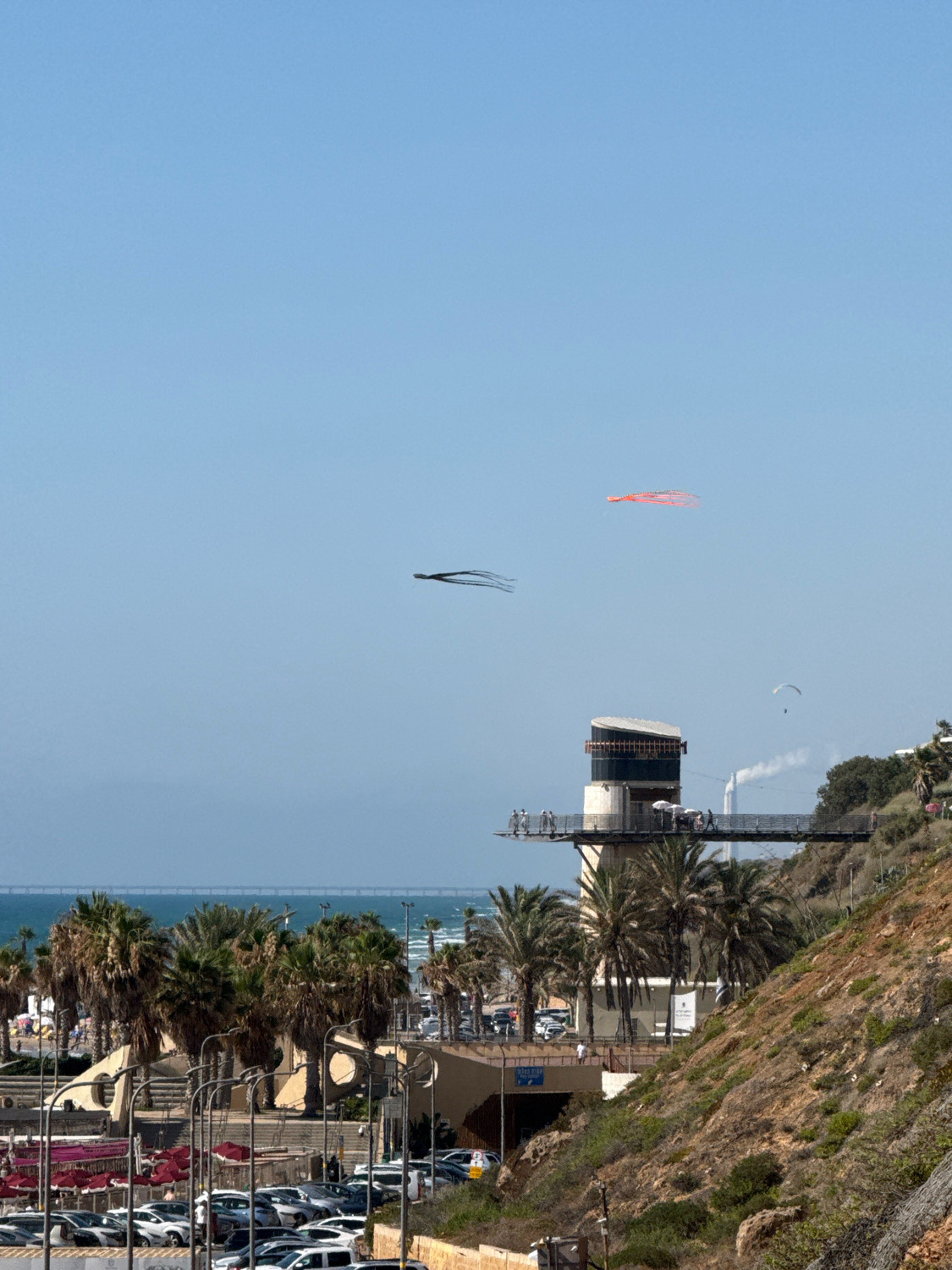The image shows a coastal scene in Netanya, Israel, with the blue sky dominating the upper portion. In the sky, there are two kites visible; one is a dark, multi-tailed kite on the left, and the other is a smaller, orange kite with a trailing tail on the upper right. Below the kites, a lookout tower with a viewing platform extends outwards from a grassy hill. Several people are standing on the platform, and a paraglider is visible in the distance, high in the sky above the tower. A plume of white smoke rises from a structure behind the tower. In the mid-ground, palm trees line a walkway leading to the tower. Further down, a parking lot is filled with numerous cars. Beyond the trees and parking lot, the sea is visible with a long bridge or pier stretching across it in the distance. The foreground shows a hillside covered in green vegetation.