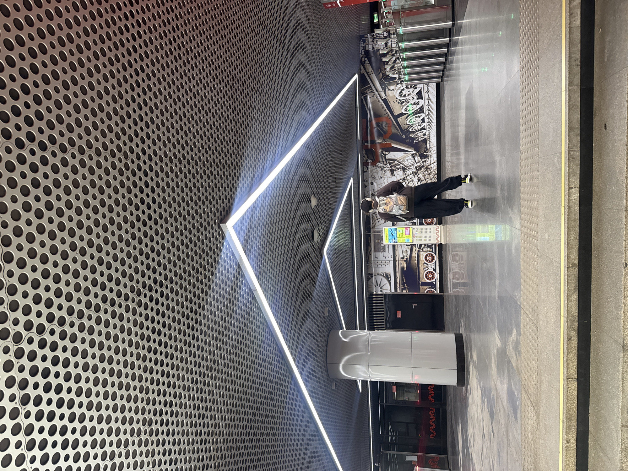 A lone person wearing a brown hoodie, black pants, and a backpack walks through a modern metro station in Moscow, Russia. The station features a distinctive perforated metal ceiling with angular white LED lighting, and a large pillar. The walls are adorned with a mural depicting mechanical elements, possibly trains or industrial machinery. In the background, automated turnstiles are visible. The floor is a polished grey material that reflects the lights and the surroundings. The image captures a typical scene in an urban transportation hub.
