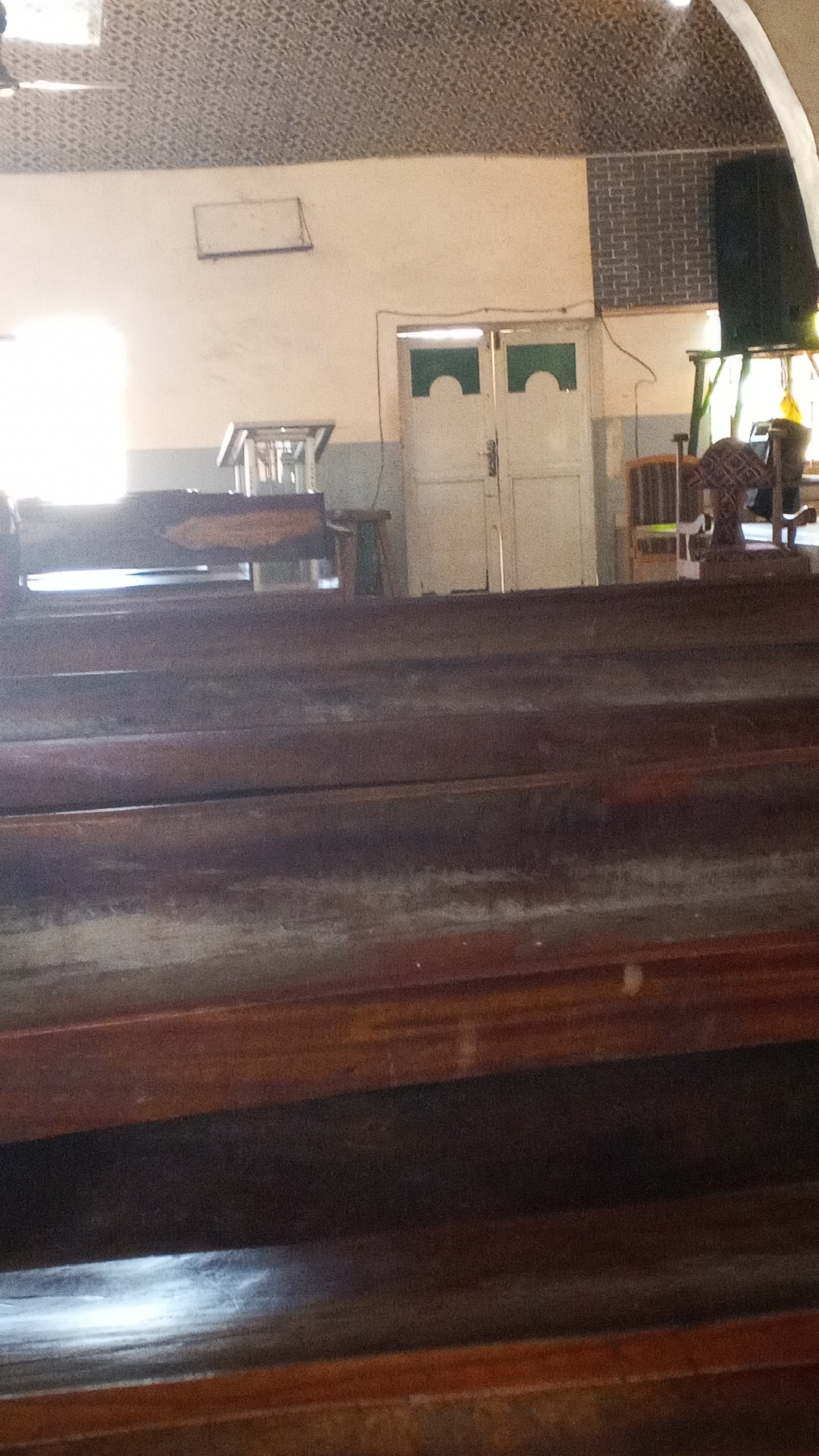 The image captures the interior of what appears to be a building, possibly a church or assembly hall, with a strong focus on wooden seating in the foreground. The seating consists of several horizontal wooden planks, worn and showing signs of age, arranged in tiers. The perspective is from a low angle, looking upwards towards the rest of the interior.
In the background, a doorway with two white doors is visible. Each door has a decorative, arched cutout at the top, revealing a green interior. To the left of the doors, a white wall is illuminated by bright light coming from an unseen source, possibly a window. A rectangular object, a light-colored plaque or frame, is mounted on the wall above this bright area.
Further back, to the right of the doorway, a dark rectangular object, likely a speaker, is mounted high on a wall that features a brick-like texture. Below the speaker and to the right of the doors, a wooden chair with a patterned seat is partially visible. To the left of the doorway, a metallic structure resembling a display stand or table is present.
The ceiling appears to be textured, possibly with a patterned wallpaper or acoustic tiles. The overall lighting suggests daytime, with the bright window area casting a strong illumination. The image is taken from within the seating area, looking towards the front of the space.