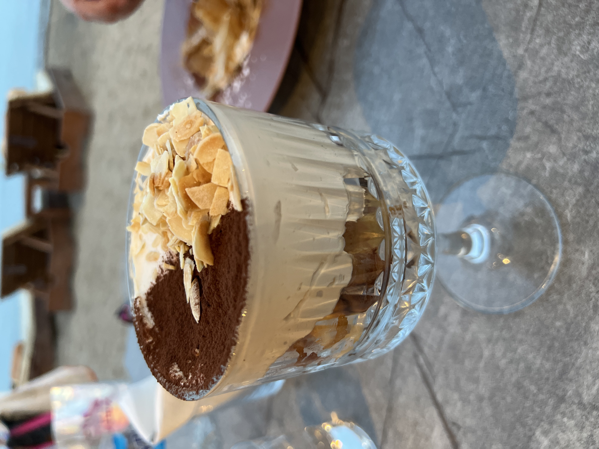 A close-up, eye-level shot captures a serving of tiramisu in a clear, fluted glass goblet. The tiramisu is layered with coffee-soaked ladyfingers and a creamy mascarpone mixture, topped with a dusting of cocoa powder and slivered almonds. The goblet rests on a gray, textured surface, possibly a stone table. In the blurred background, beach chairs and the sea are visible, suggesting an outdoor dining setting in Eretria Municipal Unit, Greece, likely a beachfront cafe or restaurant.