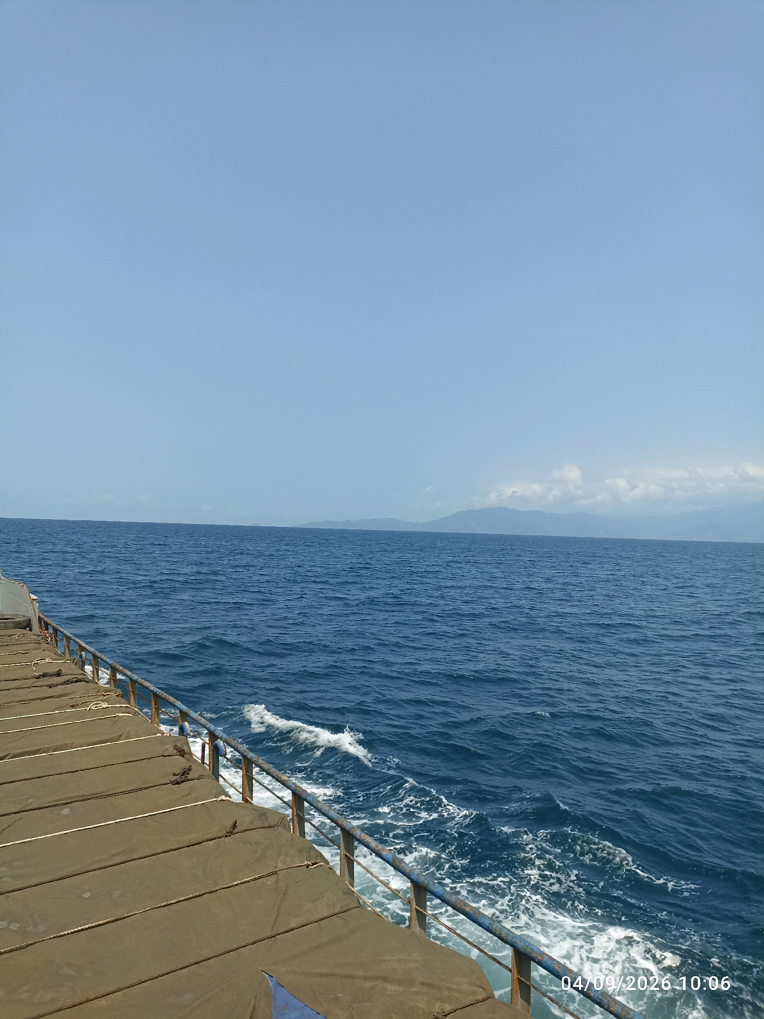 This is a photograph taken from the deck of a ferry at sea. The foreground shows the deck covered with brown tarps, secured by ropes, and a metal railing. The sea is a deep blue with white-capped waves. In the distance, a faint silhouette of mountains and a cloudy sky can be seen. The date and time stamp "04/09/2026 10:06" is visible in the bottom right corner. The image appears to be taken in Indonesia, though the specific city is unknown.