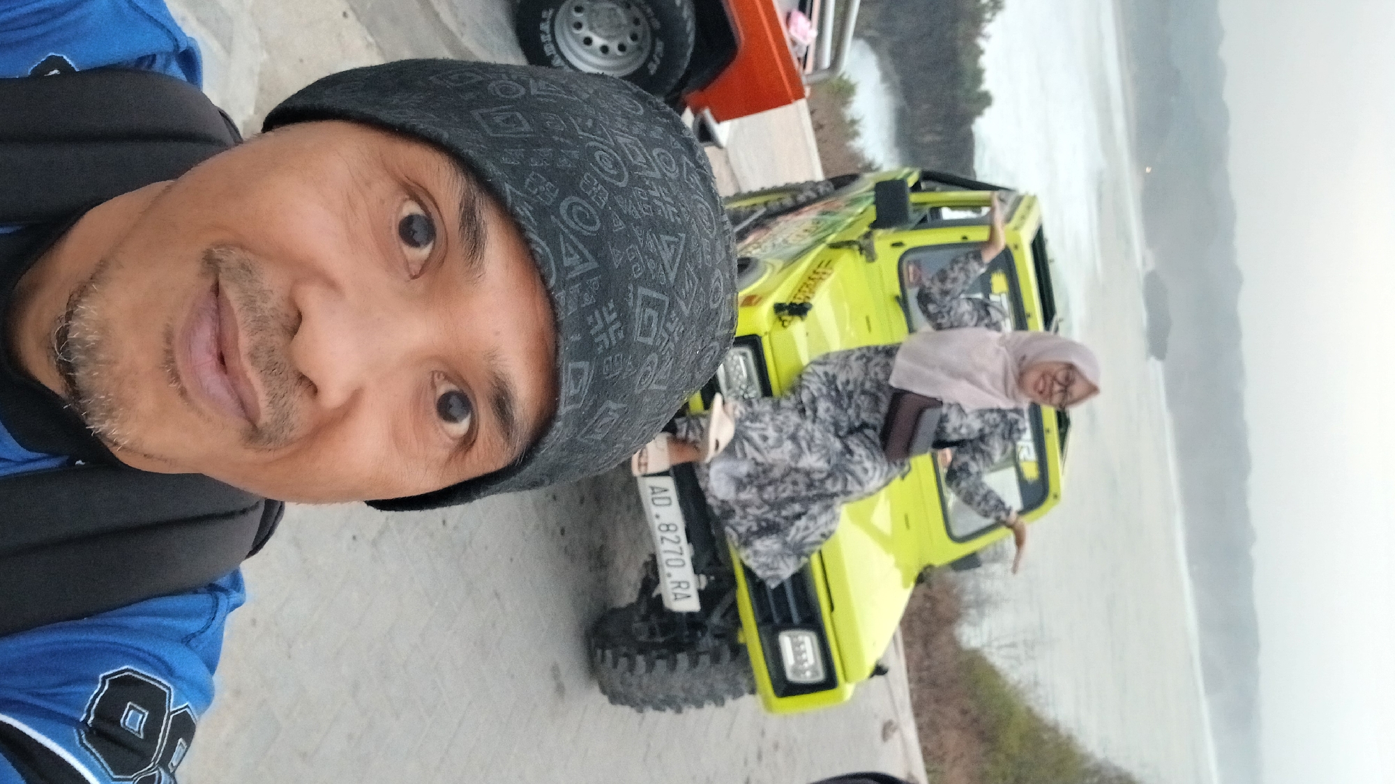 In Tanjungsari, Indonesia, a man in a black beanie and blue jacket is taking a selfie. Behind him, a woman in a hijab and a patterned dress is sitting on the hood of a bright yellow jeep, with a brown bag slung across her body. The jeep has a license plate that reads "AD.8270.RA." In the background, the ocean meets a hazy coastline with rolling hills. Another vehicle, partially visible and orange, is to the left of the jeep. The ground appears to be paved.