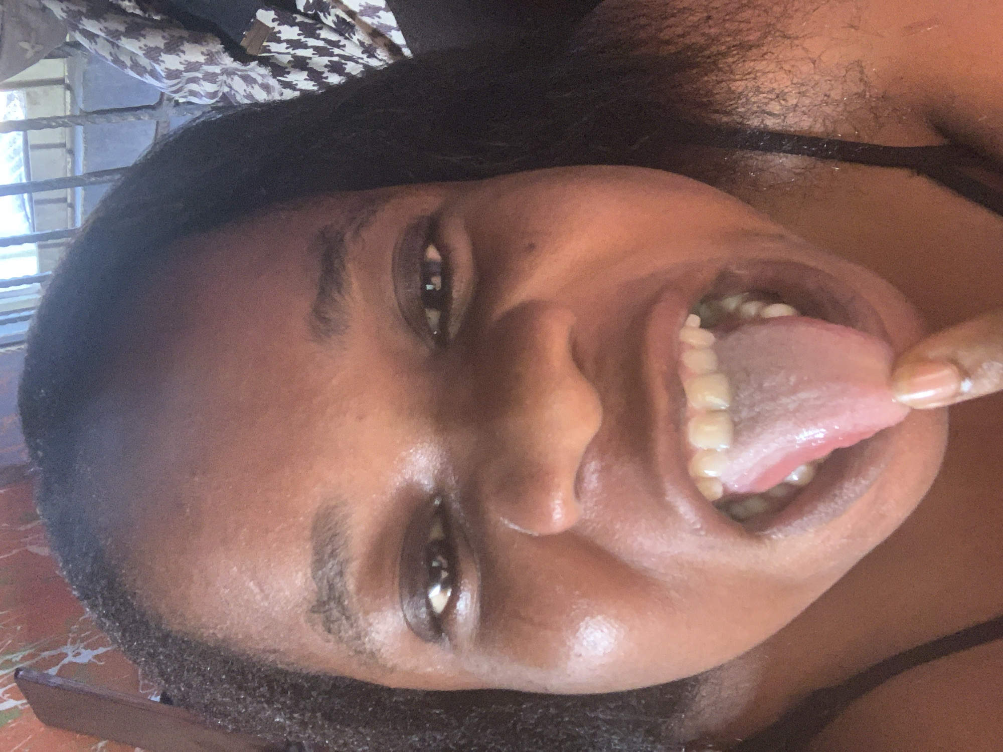 A close-up, angled, selfie-style image depicts a Black woman with brown eyes, her mouth open, tongue extended and grasped with her left hand. She has dark hair and is wearing a black strap top. The background is indistinct but includes what appears to be a window with a metal grate and a patterned cloth. The location in the city is Unknown, Unknown.