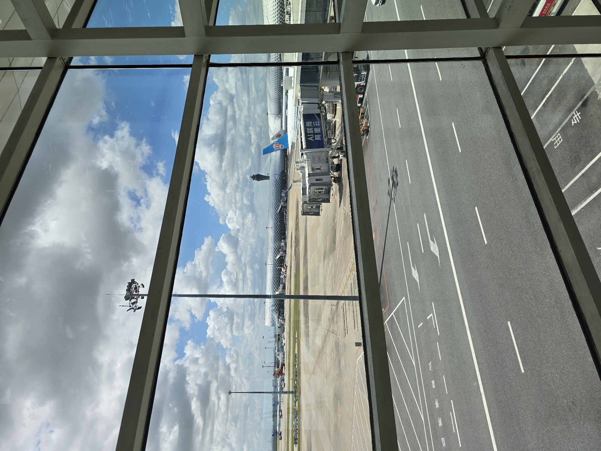 The image shows an airport tarmac viewed through a large glass window. In the foreground, a gray asphalt road is marked with white lines, indicating lanes and parking spaces. Beyond the road, the airport tarmac stretches out, with various airplanes visible, including a large white passenger jet with a blue tail logo, connected to a jet bridge. Several other aircraft are parked in the distance. The background features a modern airport terminal building with a distinctive curved, patterned roof. A tall control tower is visible to the left of the terminal. The sky is partly cloudy with a mix of blue sky and white cumulus clouds. The scene appears to be a sunny day at an international airport. The location is identified as Bao'an District, China.