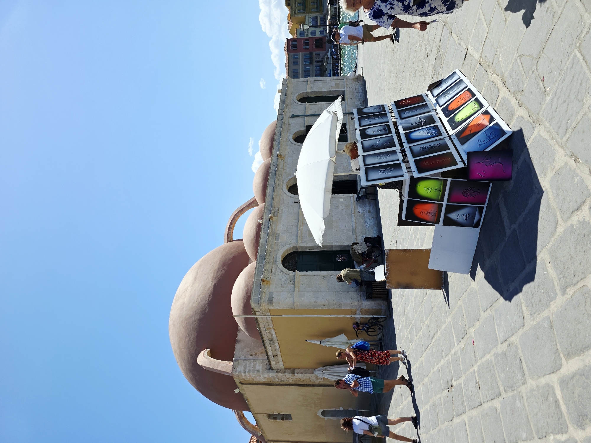 The image depicts the historic Etz Hayyim Synagogue in Chania, Greece, on a sunny day. A large, reddish-brown dome dominates the upper left portion of the frame, with smaller domes visible behind it. The building's exterior features pale stone walls and arched windows. In front of the building, to the right of center, a display of framed artwork is set up under a white umbrella, featuring colorful spray-painted images and stencils. Several people are present in the scene. On the left, a woman in a white t-shirt and shorts walks away from the camera, and a couple in checkered shirts and shorts, and a floral dress respectively, walk towards the building. A person in a green shirt is near the entrance of the building, appearing to interact with the display. Further to the right, a man in a white t-shirt and shorts walks along the waterfront, and a woman in a blue and white floral dress walks towards the foreground. The ground is paved with large, grey cobblestones. In the background, colorful buildings line the harbor, and the sea is visible. The sky is a clear, bright blue.