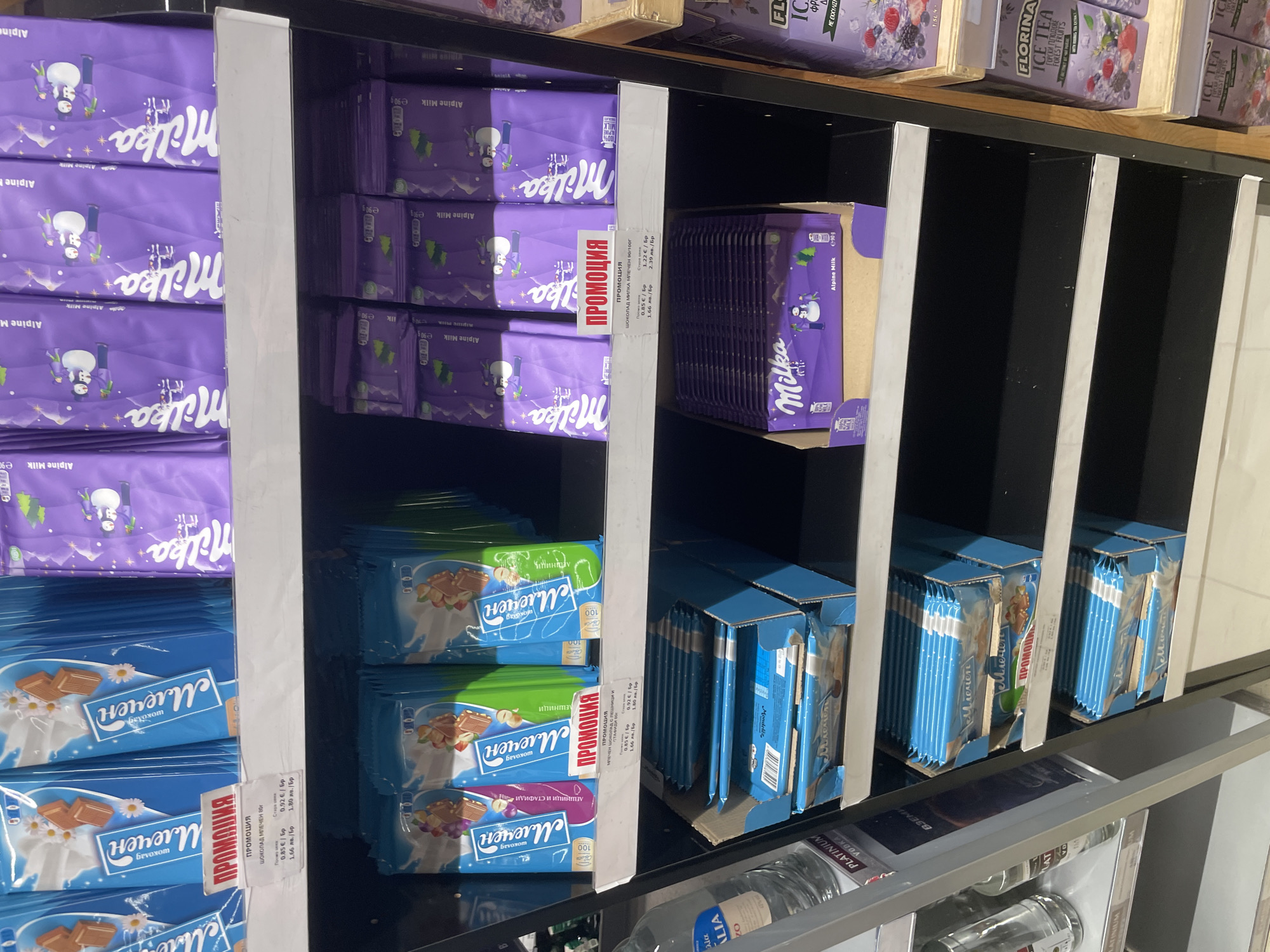 The image displays a well-stocked section of supermarket shelves in Ihtiman, Bulgaria, featuring chocolates, ice tea, and vodka. The products are organized in multiple vertical rows within metal shelving units with white dividers.
The left side of the frame is dominated by Milka Alpine Milk chocolate bars, identifiable by their purple packaging which features a snowman, snowy mountains, and cows. These 90g bars are shown stacked and within a cardboard display box. A red "ПРОМОЦИЯ" (Promotion) label indicates a discounted price of 0.85 € (1.66 лв.) per bar, down from 1.22 € (2.39 лв.).
Below the Milka chocolates, and extending to the right, are "Млечен" (Milk) brand chocolate bars. These are available in blue, green, and purple packaging, offering varieties like plain milk chocolate, hazelnuts ("ЛЕШНИЦИ"), and hazelnuts with raisins ("ЛЕШНИЦИ И СТАФИДИ"). Similar "ПРОМОЦИЯ" labels are present for these chocolates, showing a promotional price of 0.85 € (1.66 лв.) from an original price of 0.92 € (1.80 лв.) for the 80g bars. Some Mlechen chocolates are stacked, while others are arranged in blue cardboard display boxes.
At the top right, bottles of Florina Ice Tea with "ГОРСКИ ПЛОДОВЕ" (Forest Fruits) flavor are visible. Towards the bottom right of the image, bottles of "PLATINIUM VODKA" from the "ALIA" brand are displayed, with the text "ВЗЕМИ" (Take/Get) partially visible.
The visible text, including brand names, product descriptions, and pricing information, is predominantly in Bulgarian, reinforcing the specified location context of Ihtiman, Bulgaria. The scene captures a typical retail display with active promotional sales.