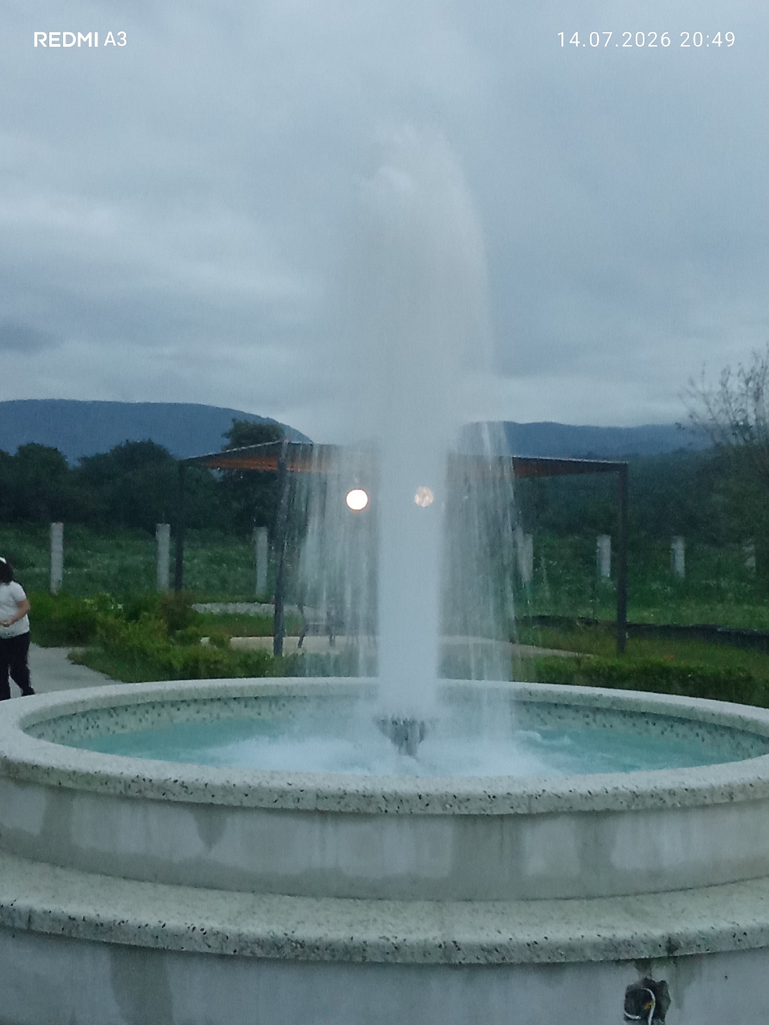 The image shows a fountain in the foreground, with water spraying upwards. The fountain has a textured, speckled concrete basin. In the background, there is a person standing on the left, wearing dark pants and a light-colored top. Beyond the fountain and the person, there is a grassy area with some bushes and trees. In the far background, a line of hills or mountains is visible under a cloudy sky. The scene appears to be outdoors in a park or garden setting. The text "REDMI A3" is in the top left corner, and "14.07.2026 20:49" is in the top right corner, indicating the device used and the date and time the photo was taken. There is no information in the image to confirm the location as Markhiaul, Abkhazia.