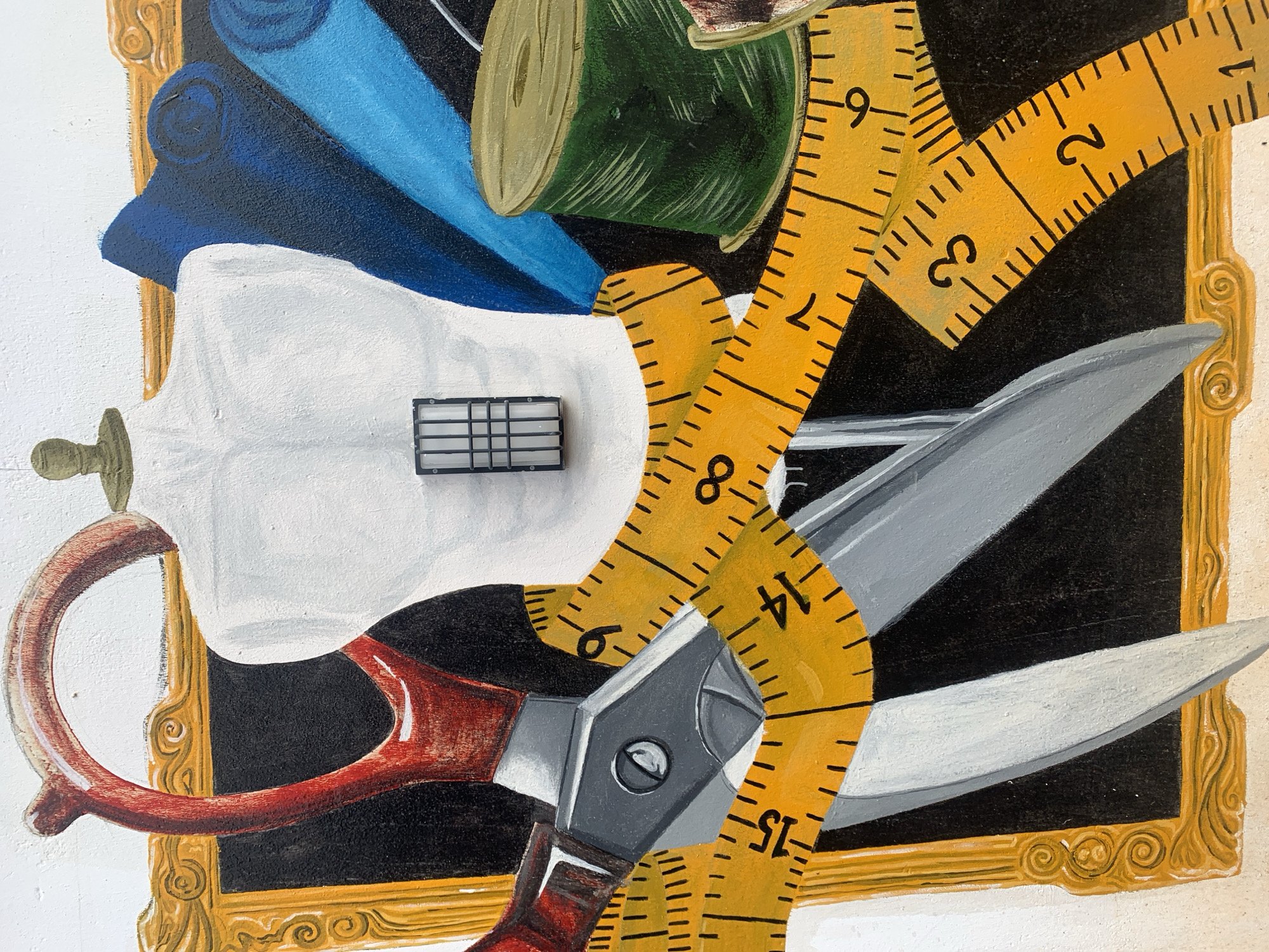 This is a close-up, detailed photograph of a painting, likely a mural, depicting sewing supplies. In the center, a white tailor's dummy is visible, with a small, black, grid-like object attached to its chest. Wrapped around the dummy and extending into the foreground is a bright yellow measuring tape marked with black numbers and lines, indicating inches or centimeters. To the left of the dummy are large, red-handled scissors, their metallic blades open. Behind the dummy, to the right, are rolls of fabric, one a deep blue and another a lighter blue. A spool of green thread is also prominent. The background is mostly black, but framed by an ornate gold border on the left and partially on the top and right. The style of the painting is realistic with bold brushstrokes. The image appears to be an outdoor painting on a textured surface, suggesting it is a mural on a wall in Gura Top, Nigeria. There are no people or animals depicted in the scene.