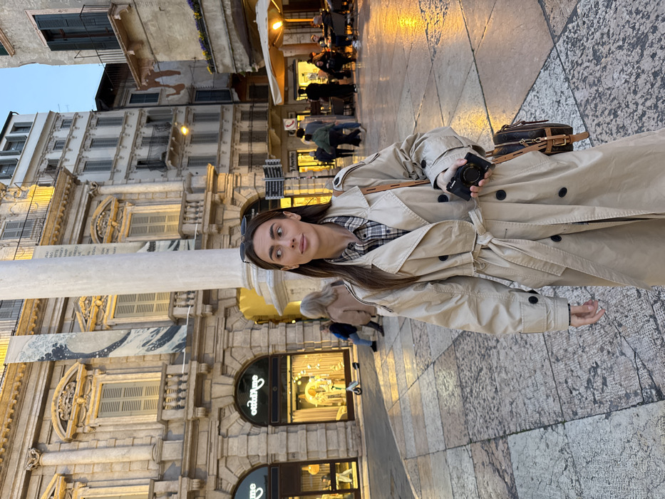 A light-skinned female, appearing to be in her twenties or thirties, with long brown hair, stands in the Piazza delle Erbe, Verona, Italy. She wears a light tan double-breasted trench coat with a belted waist over a plaid shirt, and sunglasses are perched on her head. She holds a small black digital camera in her left hand, and a brown patterned shoulder bag with a light brown strap is on her right shoulder. Her gaze is directed to her right (viewer's left) with a neutral expression.
Behind the subject, the historical architecture of Piazza delle Erbe is visible. To the left, the ornate stone facade of the Palazzo Maffei features multiple stories, arched windows with shutters, and two vertical banners depicting "The Great Wave off Kanagawa." Ground-floor shops, including "Otticame" (an optician), are visible. A tall, fluted white marble column stands to the right of the Palazzo Maffei. To the right of the column, additional multi-story buildings display windows, some with closed shutters, and a ground-level outdoor café area with yellow artificial lighting and red awnings/umbrellas. Several indistinct figures are seated at the café tables or walking in the background. The ground is paved with irregular, light-colored stone blocks. A dark signpost with multiple directional signs is visible in the mid-ground. An electric scooter and a person sitting on a step are also present in the background to the left. The scene is illuminated by natural light, suggesting late afternoon or early evening, complemented by artificial yellow light from the establishments.