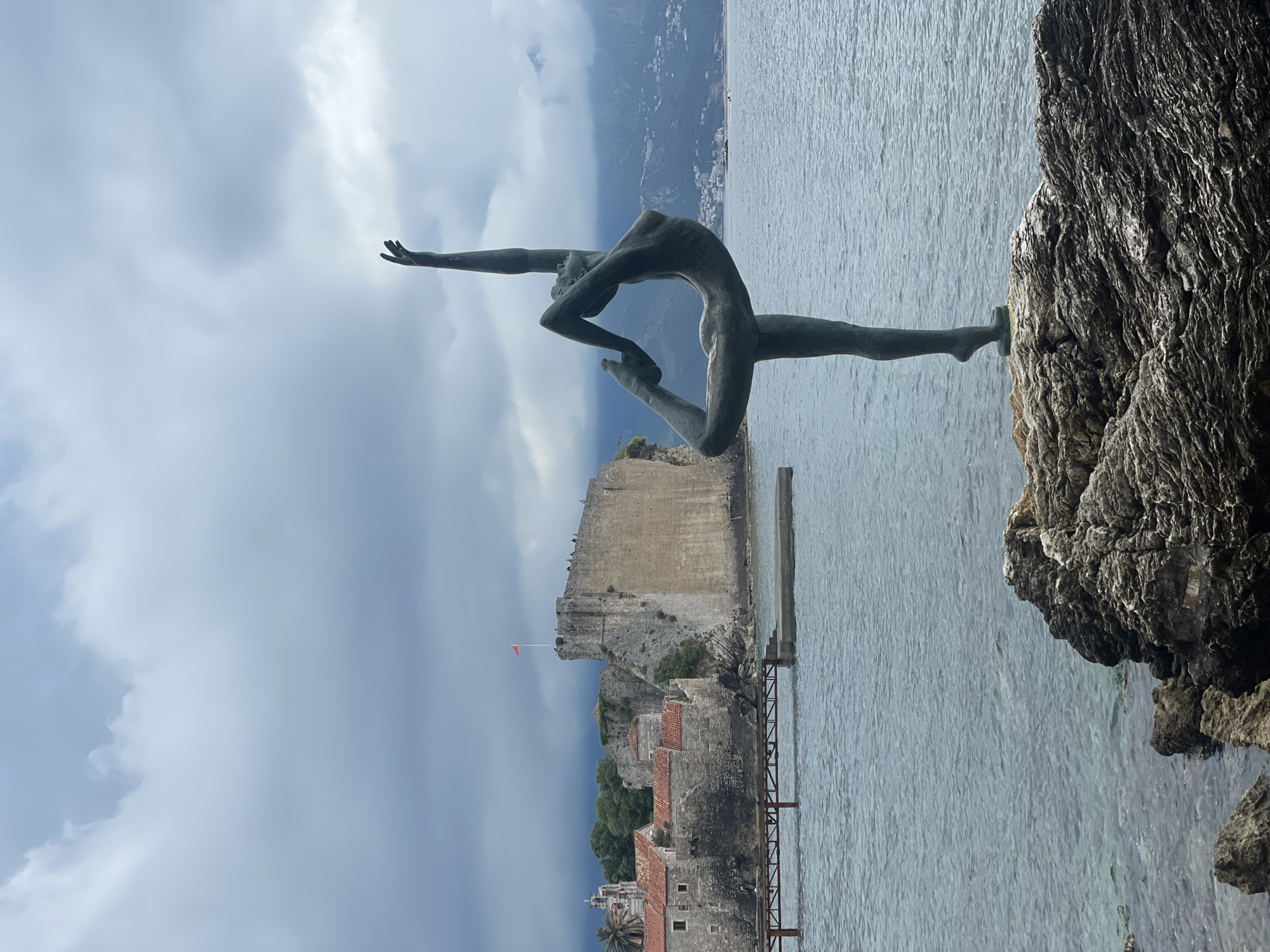 The image shows the "Ballerina of Budva" statue in Budva, Montenegro, standing on a rocky outcrop. The statue is of a woman in a dynamic yoga pose. Behind the statue, the old town of Budva is visible, along with a portion of the Adriatic Sea and distant mountains under a cloudy sky.